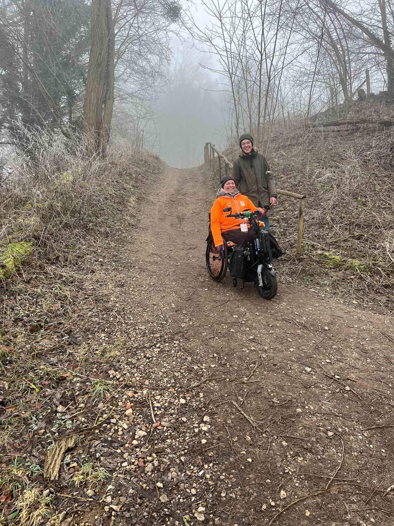 Twee mensen, één in een rolstoel, wandelen op een modderig bospad met mistige achtergrond en kale bomen.