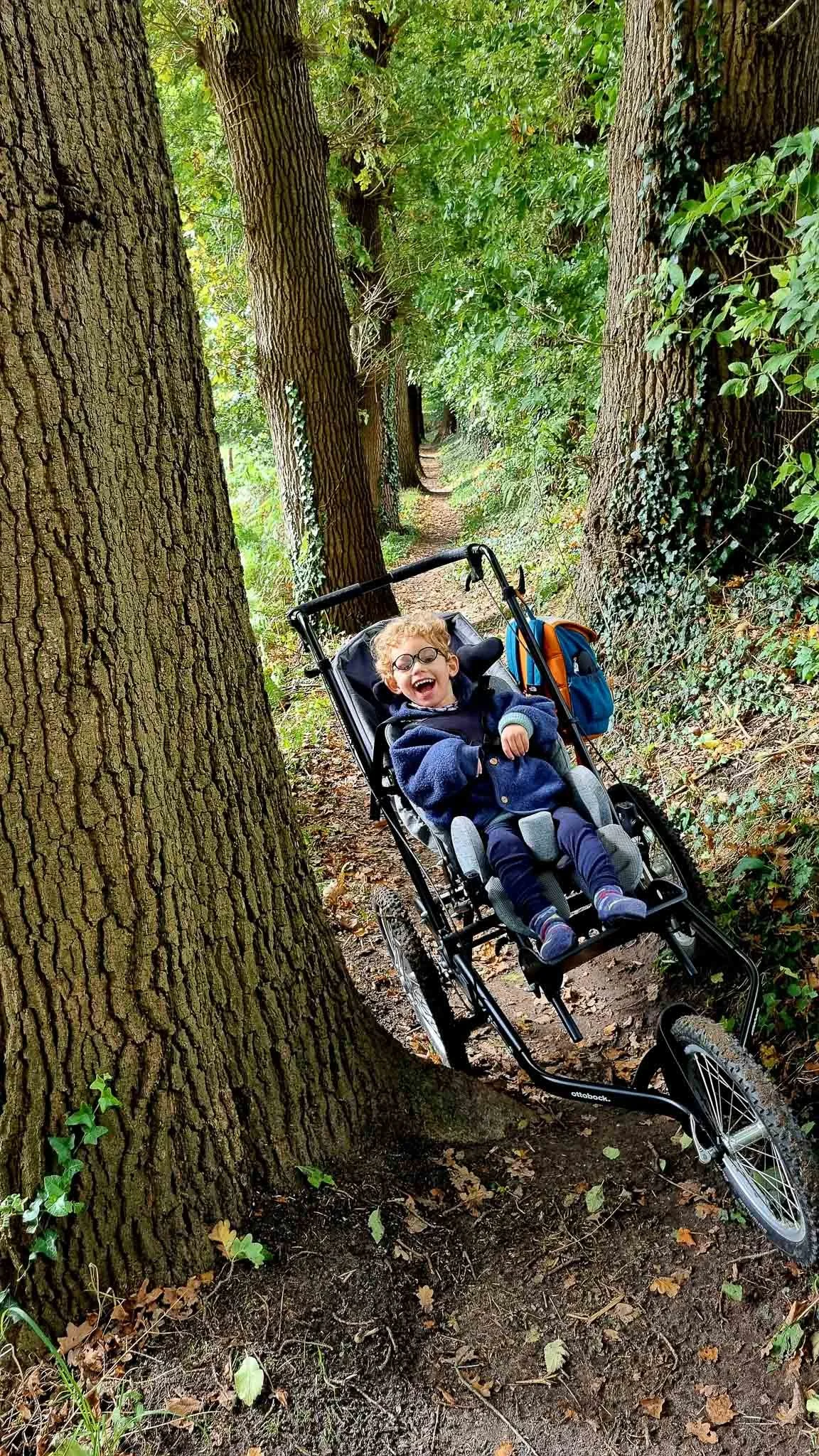 Een kind in een rolstoel fietsen door een bos met grote bomen en groene bladeren.
