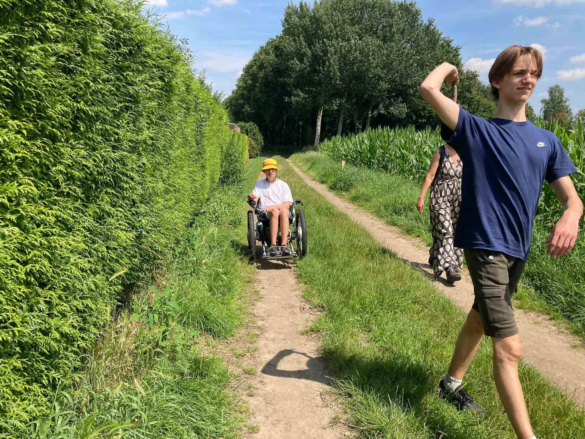 Twee jonge mensen wandelen op een smal pad langs een groen veld met maisplanten, een jongen helpt iemand in een rolstoel terwijl ze genieten van het zonnige weer.