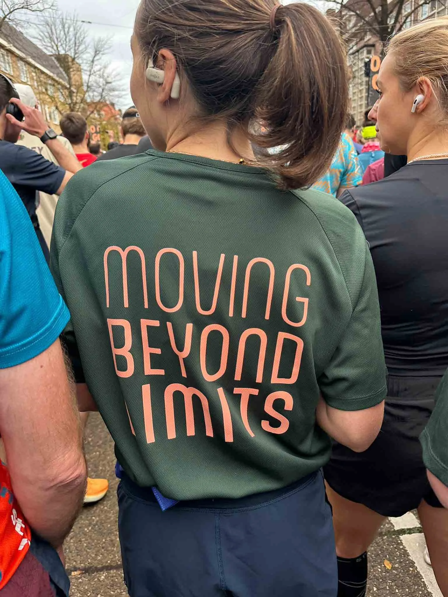 Close-up van een groen T-shirt met de tekst 'Moving Beyond Limits' in roze letters, gedragen door een persoon op een druk evenement of loopwedstrijd.