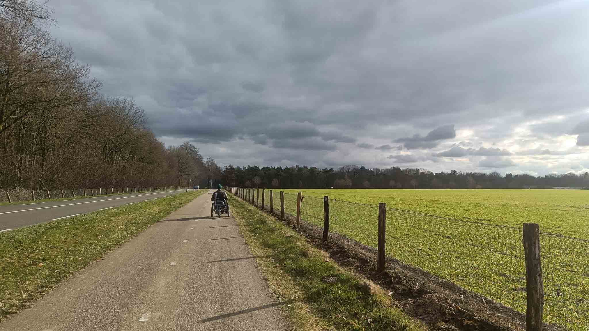 Fietser op een rustige weg naast een groen veld onder een bewolkte lucht.