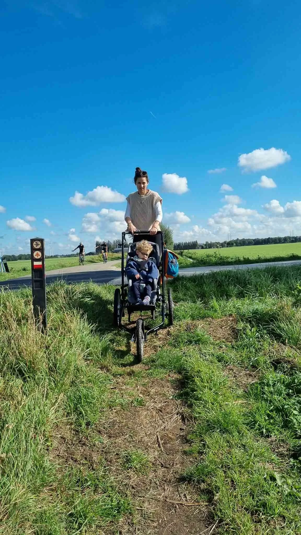 Vrouw met een kinderwagen met een kind erin, buiten op een pad in een groene omgeving met een heldere blauwe lucht.