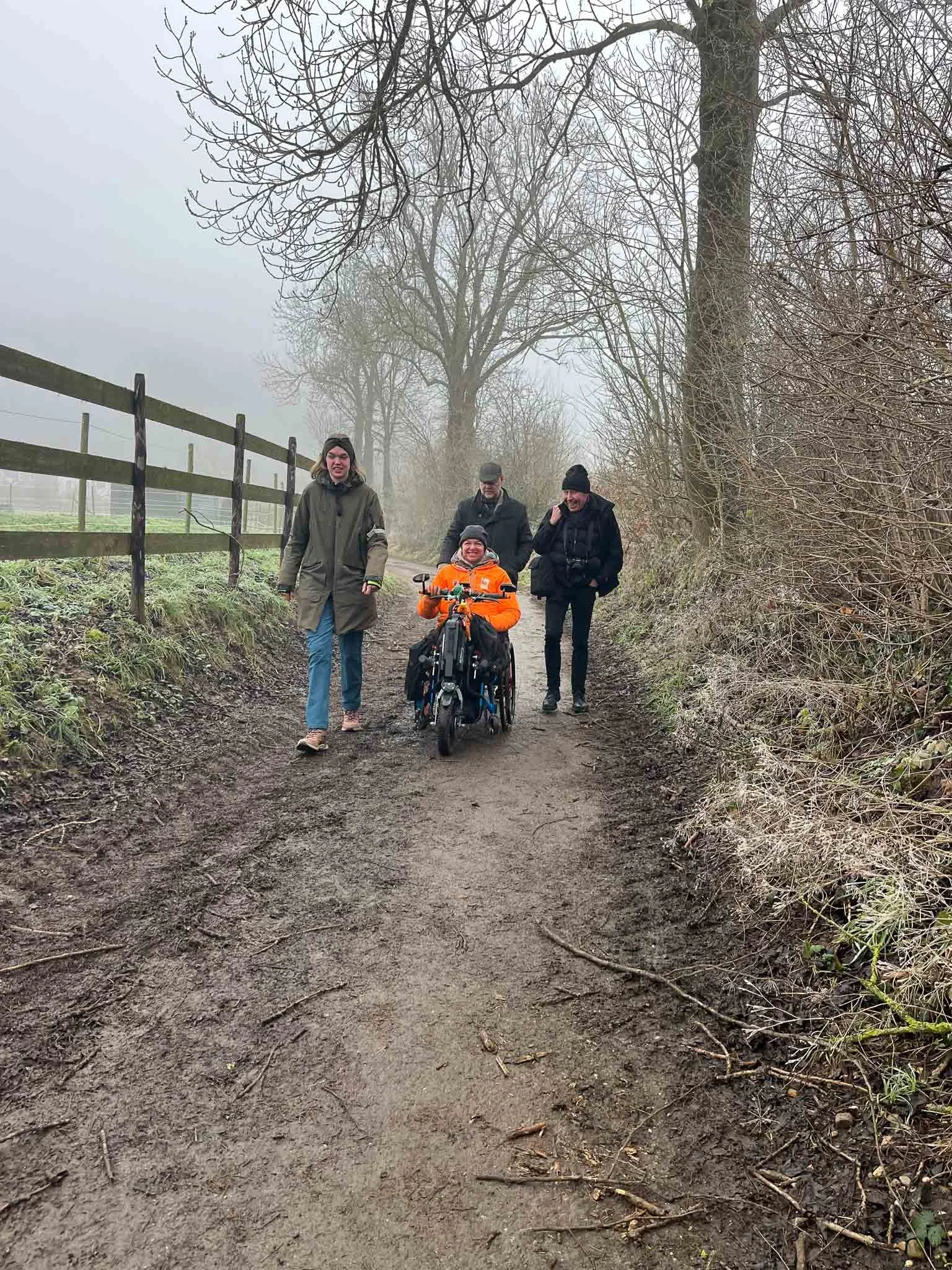 Een groep mensen wandelt op een bosweg in de winter, met mist en zonder bladeren aan de bomen, inclusief een persoon in een rolstoel.