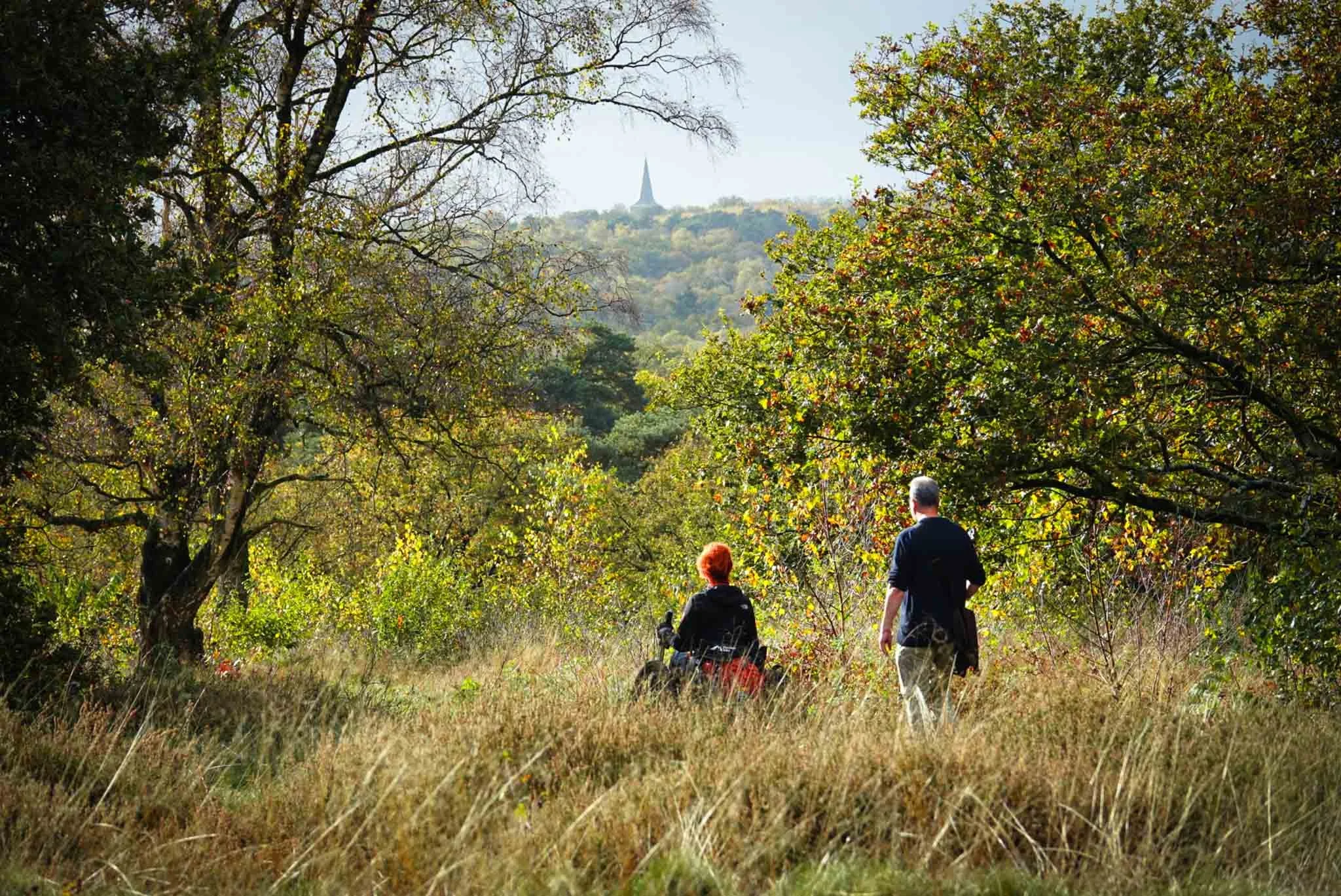 Twee mensen wandelen door een bosrijk gebied met groene bomen en gras, met een kerk of torenspits zichtbaar op de heuvel op de achtergrond.