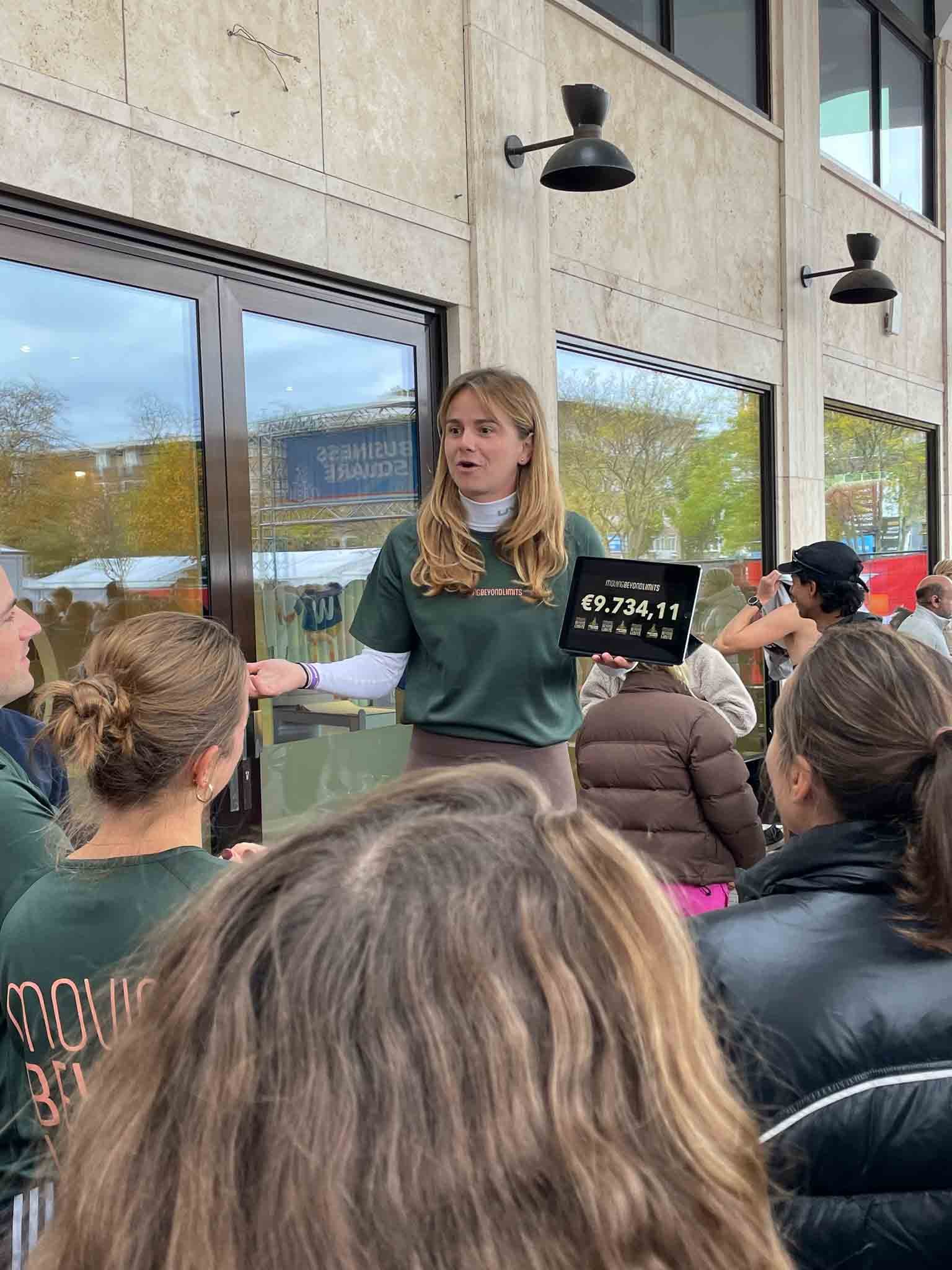 Vrouw vertelt een groep mensen buiten over een bedrag op een scherm, omgeven door mensen die luisteren en kijken.
