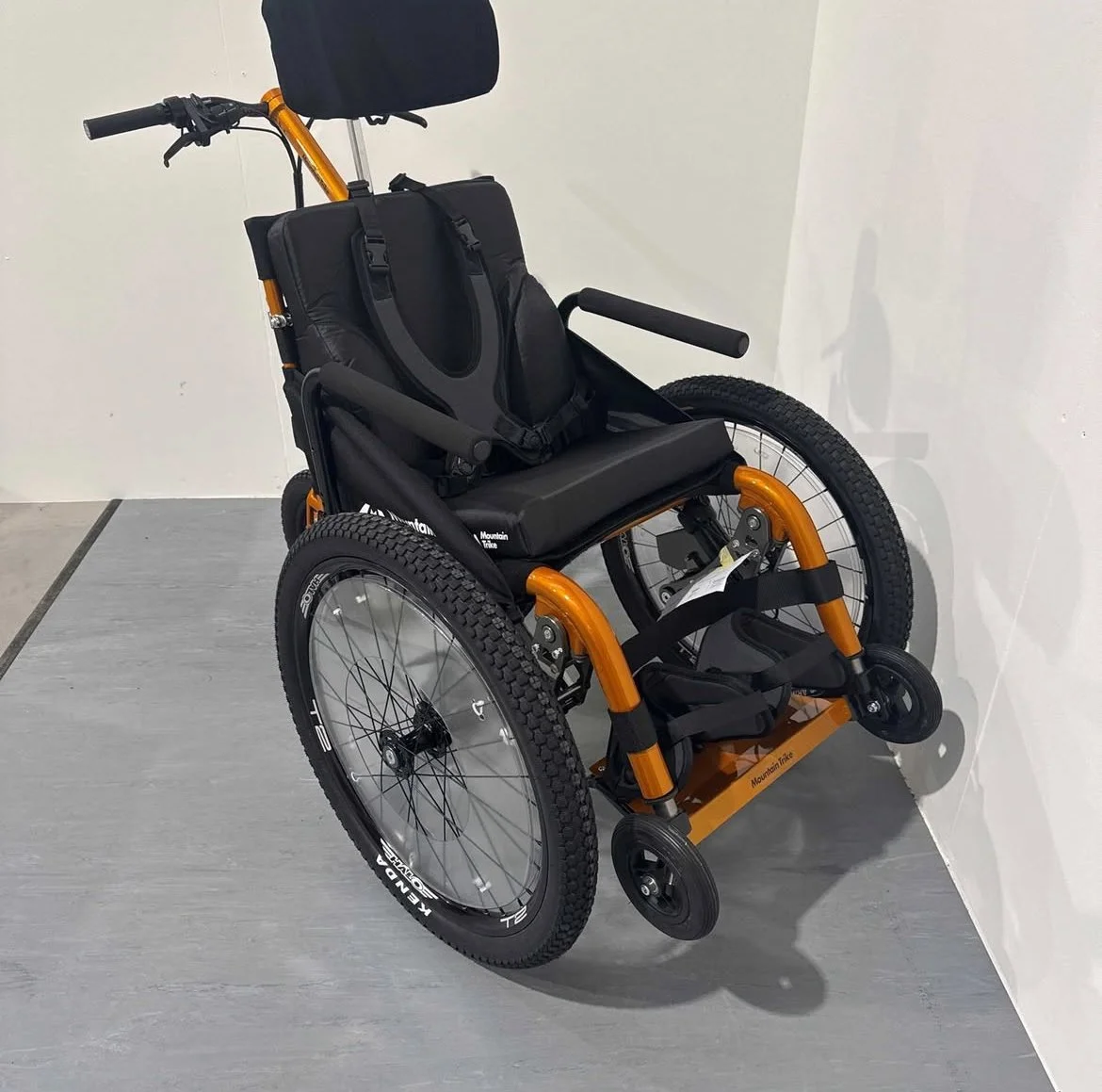 Mountain Trike e-Push. Een rolstoel met een zwart zitoppervlak, een hoofdsteun, armleuningen en grote achterwielen, die tegen een witte muur staat.