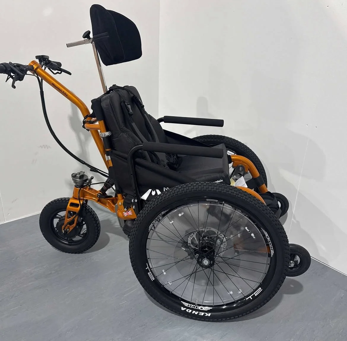 Mountain Trike e-Push. Een sportrolstoel met een houten frame en dikke banden, geplaatst in een witte kamer.