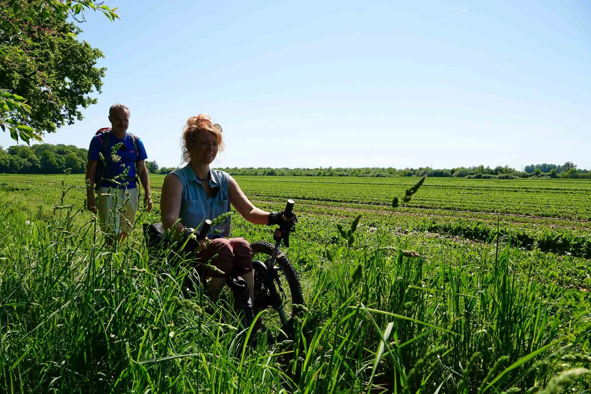 Twee mensen, een vrouw die op een mountainbike zit en een man die achter haar loopt, op een groen veld onder een heldere blauwe lucht.