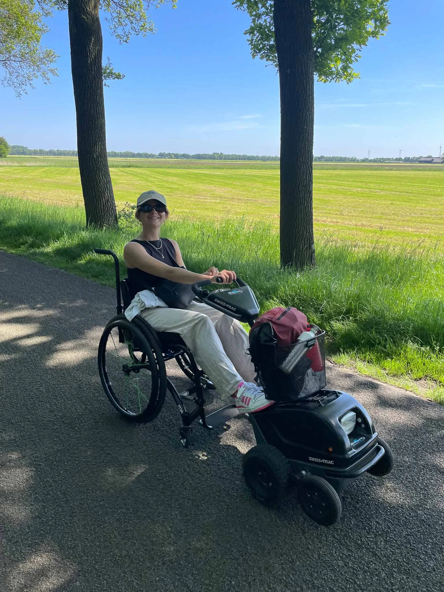 Vrouw in een rolstoel rijdt op een pad onder bomen, met een uitgestrekt grasland en blauwe hemel op de achtergrond.