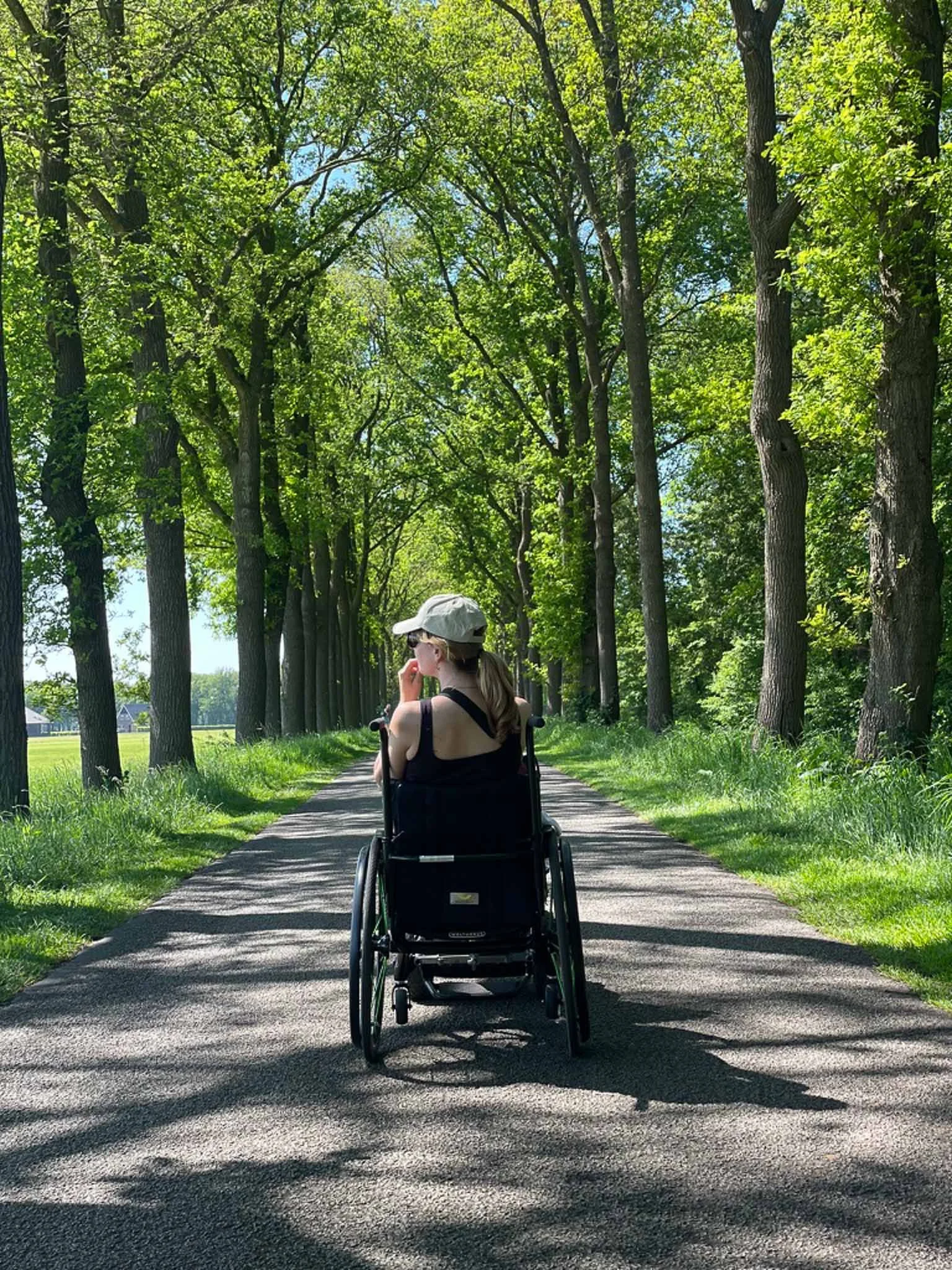 Vrouw in een rolstoel rijdt langs een tree-lined pad op een zonnige dag, omgeven door groene bladeren.