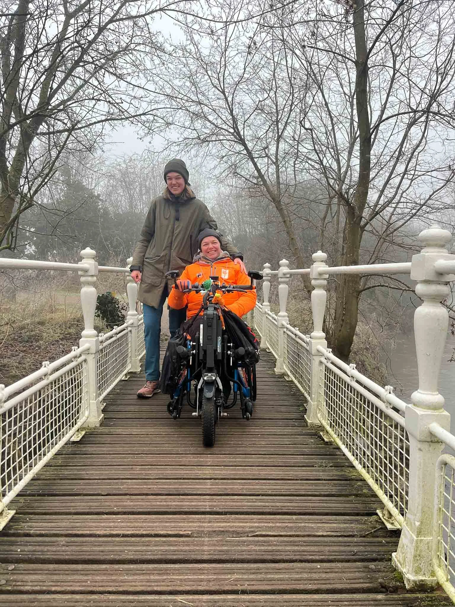 Twee vrouwen op een kleine houten loopbrug in een park, één staat en één zit in een rolstoel, omringd door kale bomen op een mistige dag.