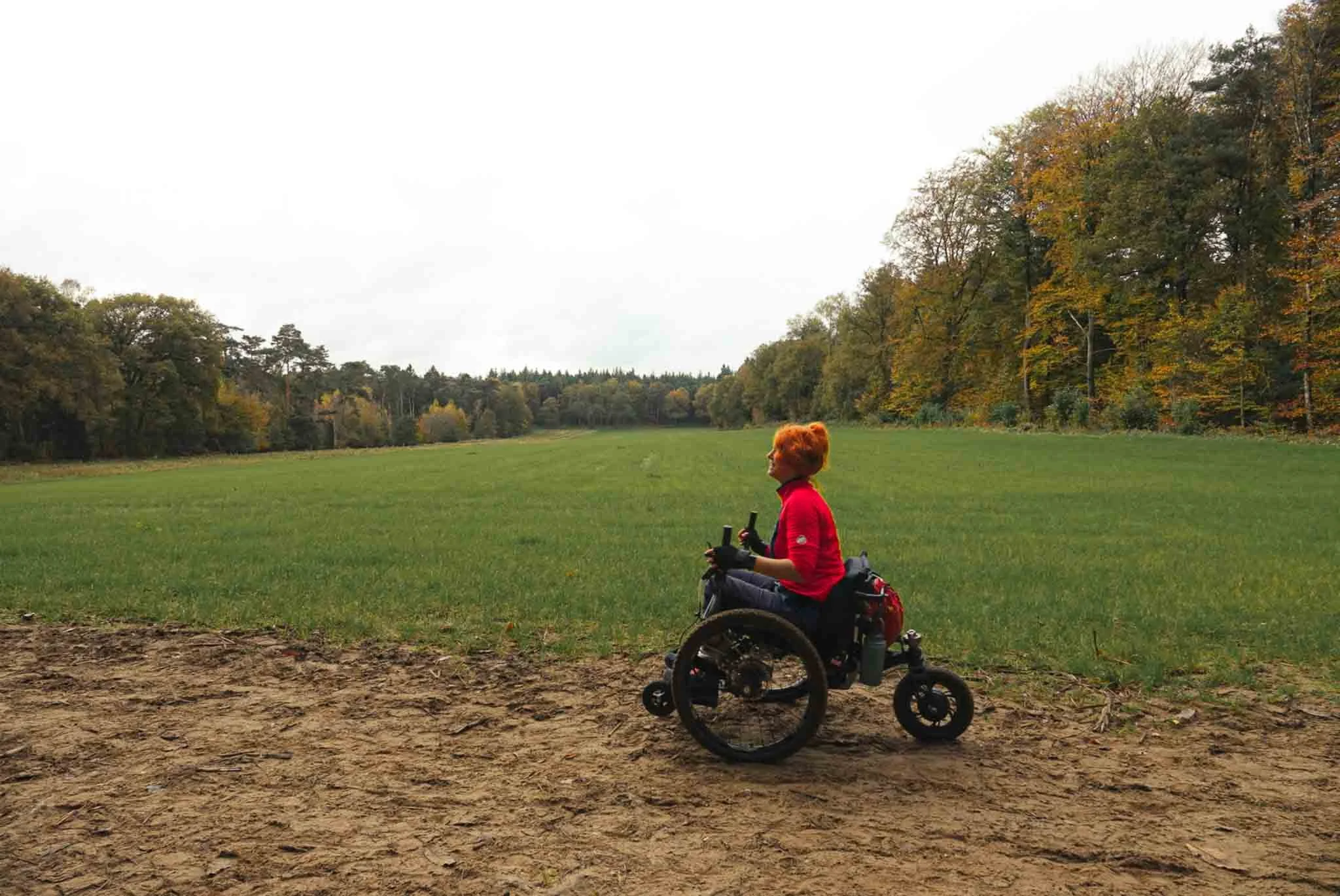 Vrouw met rood haar in een rolstoel op een pad in een groen veld met bomen in de herfst, onder een bewolkte sky.