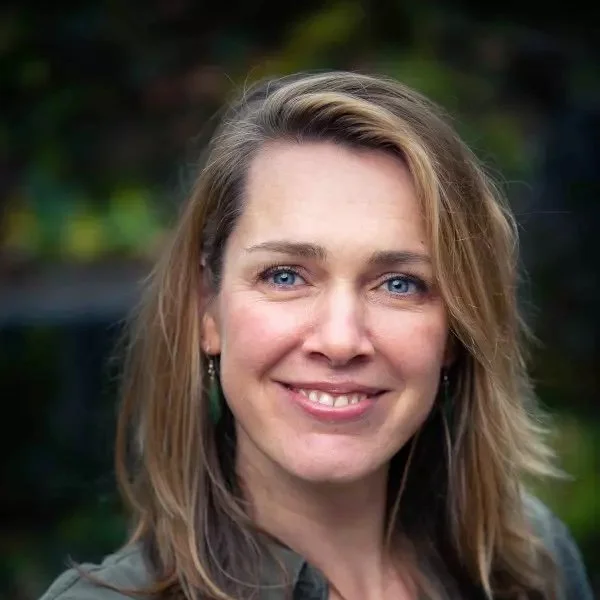 Close-up foto van een lachende vrouw met blond haar en blauwe ogen, buiten met groene achtergrond.