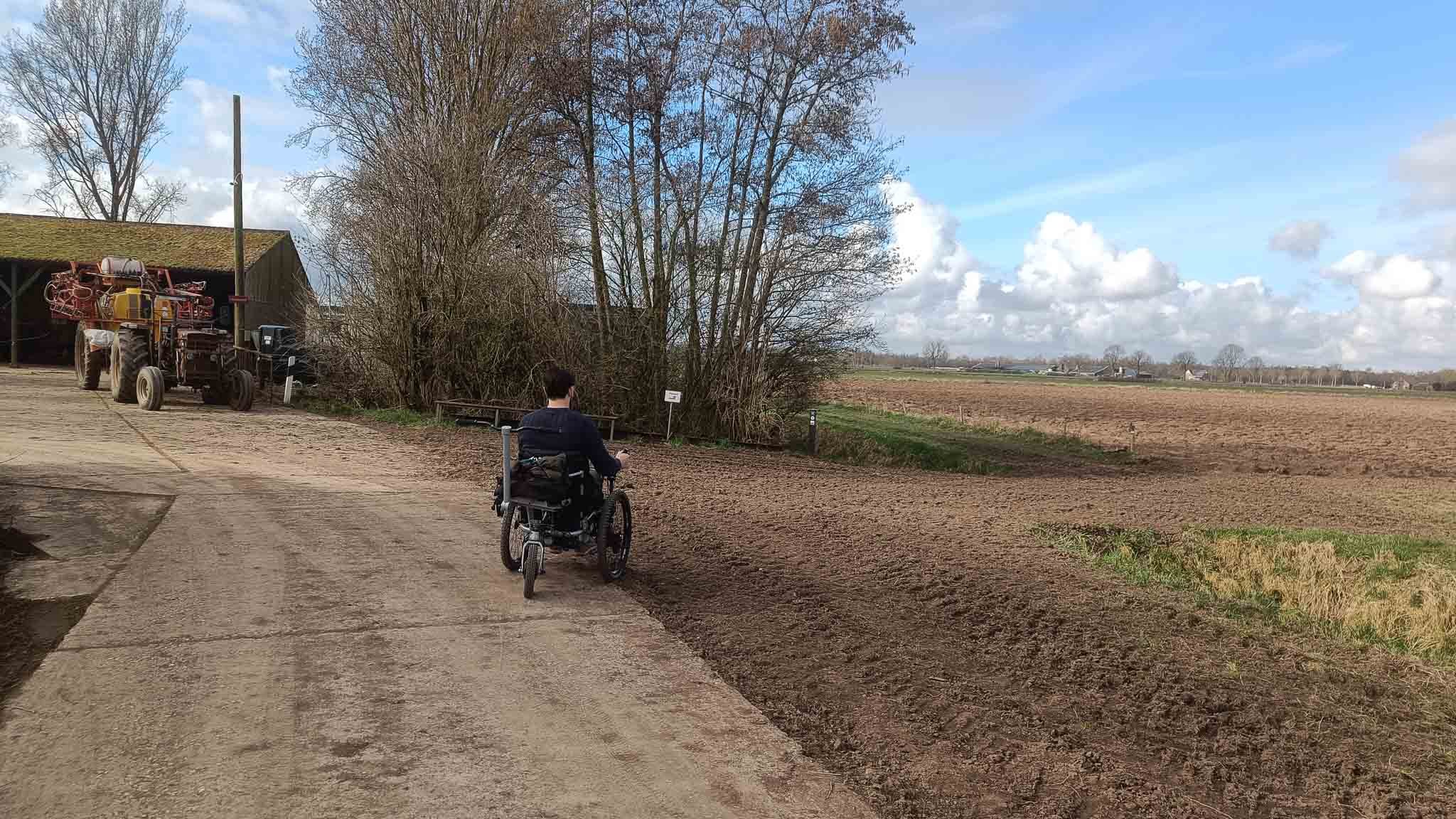 Person in een rolstoel fietst op een landweg met een veld aan de rechterkant en een boerderij met landbouwvoertuigen aan de linkerkant, onder een deels bewolkte blauwe lucht.