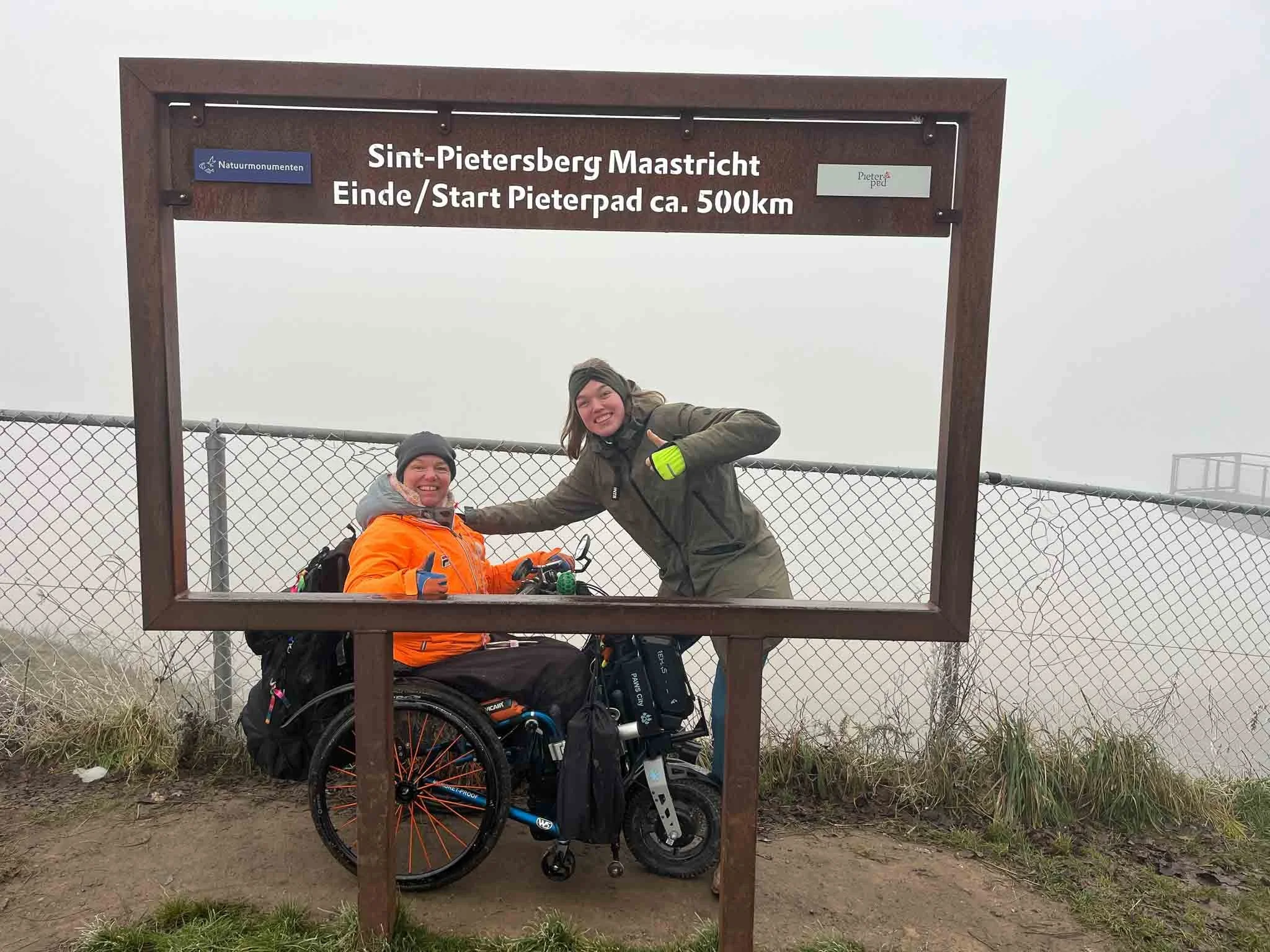 Twee vrouwen met een elektrische rolstoel bij de ingang van een bergpad, omgeven door mist, onder een houten bord met tekst over een wandelroute.