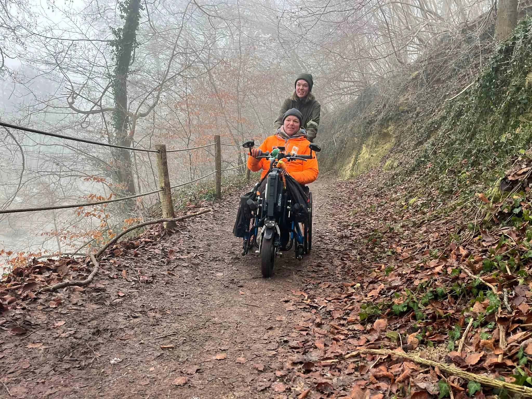 Twee mensen op een bosweg, een persoon in een rolstoel en een persoon die achter hen loopt, omgeven door bomen en struiken met herfstbladeren en mist.