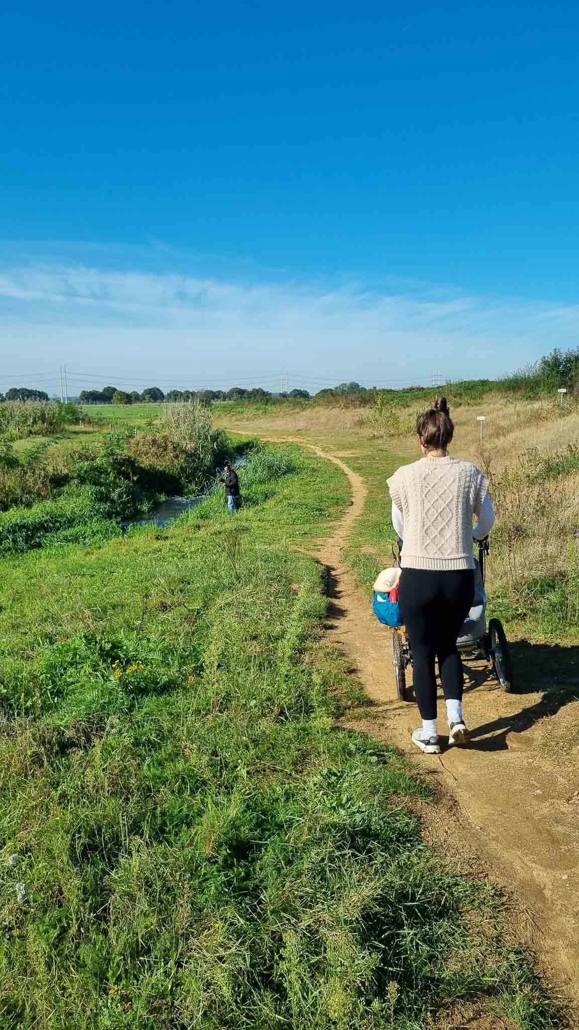 Vrouw wandelt met kinderwagen langs een pad in een groene natuur onder een blauwe lucht.