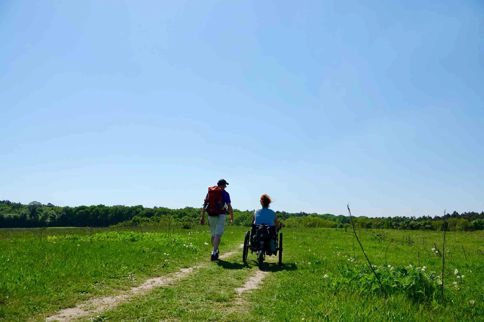 Twee mensen wandelen op een grasveld, één in een rolstoel, onder een heldere blauwe hemel.