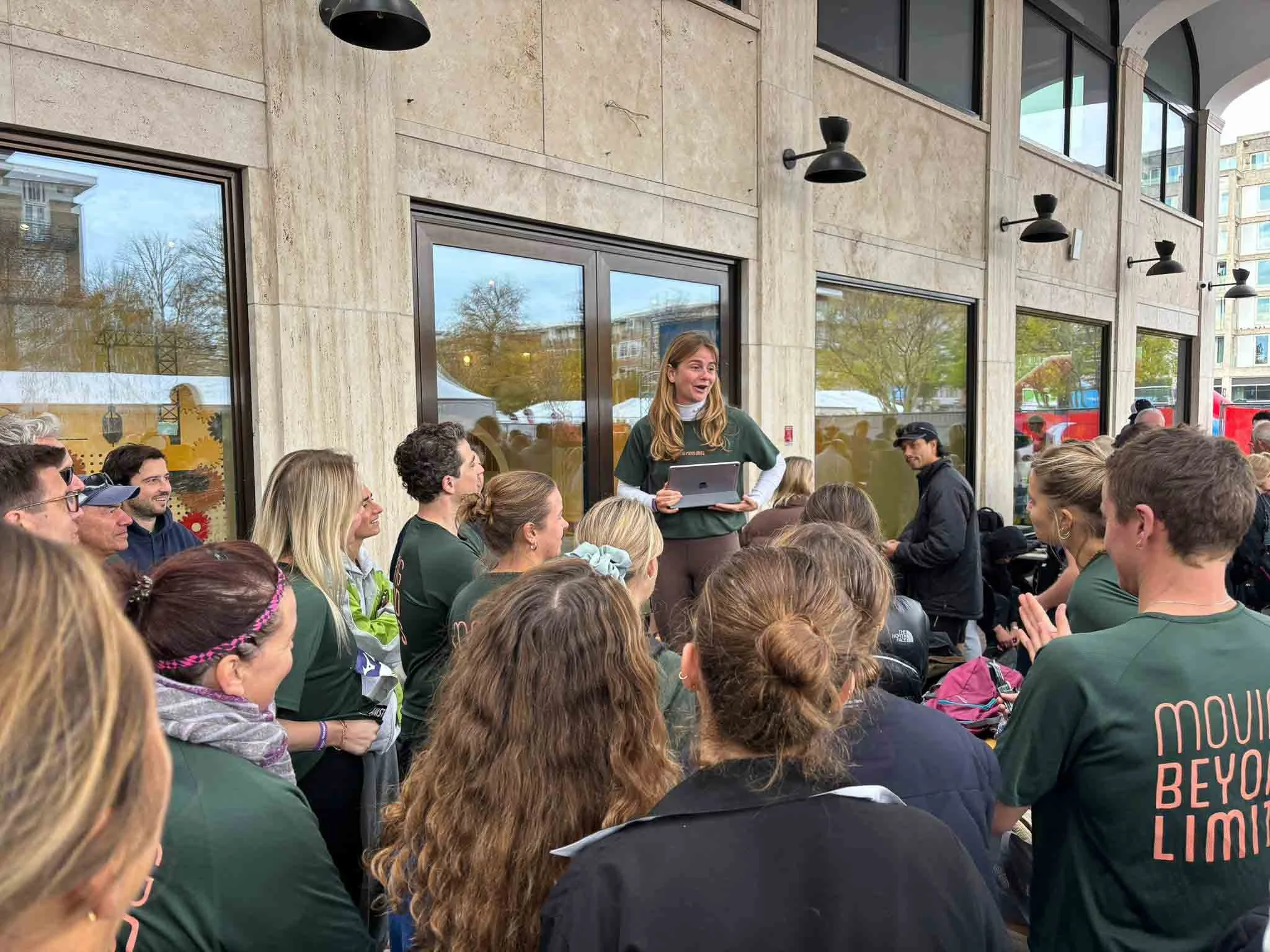 Een groep mensen die buiten zit, kijken naar een vrouw die met een tablet spreekt. Het is een grijze dag, en er is veel groen in de achtergrond.