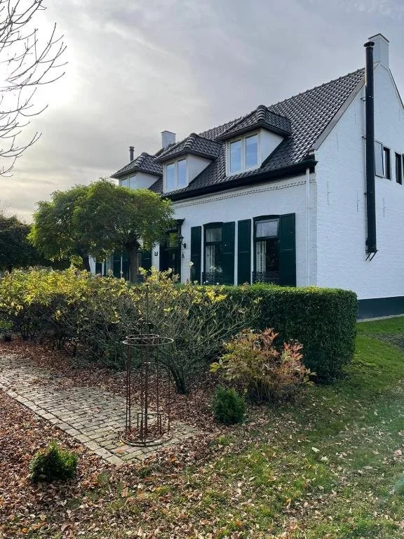 Toegankelijke accommodatie aan het Pieterpad. Een wit huis met een donkere dak, omgeven door een tuin met struiken en een boom, met een grindpad en een metalen plantenstandaard voor potten.