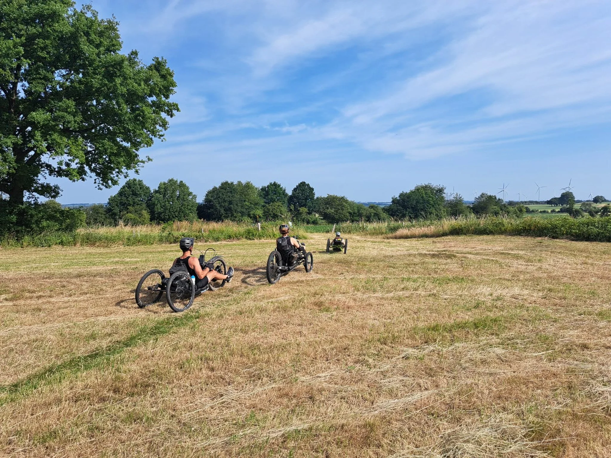 Drie mensen met rolstoelen rijden over een open veld onder een blauwe lucht met enkele wolken, omringd door groene bomen en windmolens op de achtergrond.