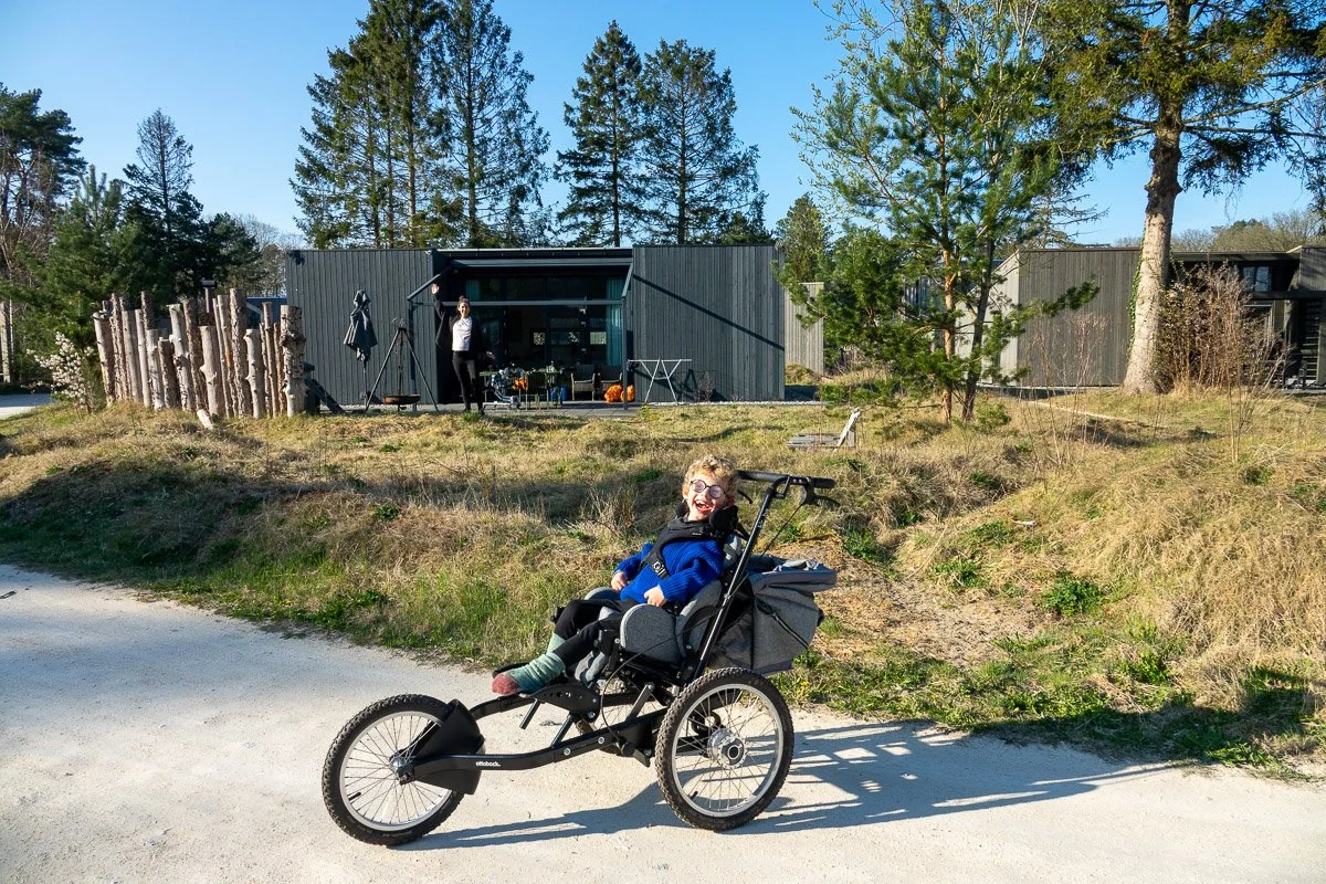 Kind in een aangepaste driewielfiets die lacht en in de zon rijdt, met een huis en bomen op de achtergrond.