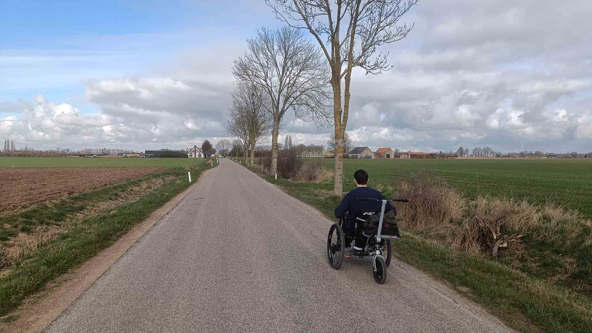 Man in een rolstoel rijdt over een smalle landweg met bomen langs de weg, groene velden en een heldere sky met wolken.