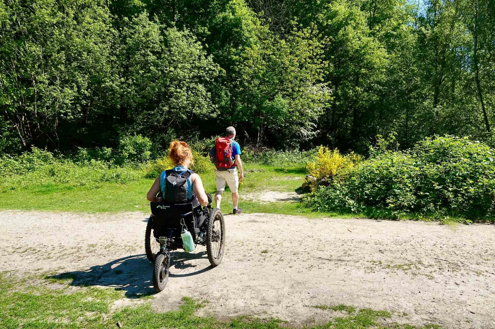 Twee mensen wandelen door een bosgebied, één persoon zit in een rolstoel en de ander loopt voor hen uit, beiden dragen backpacks.
