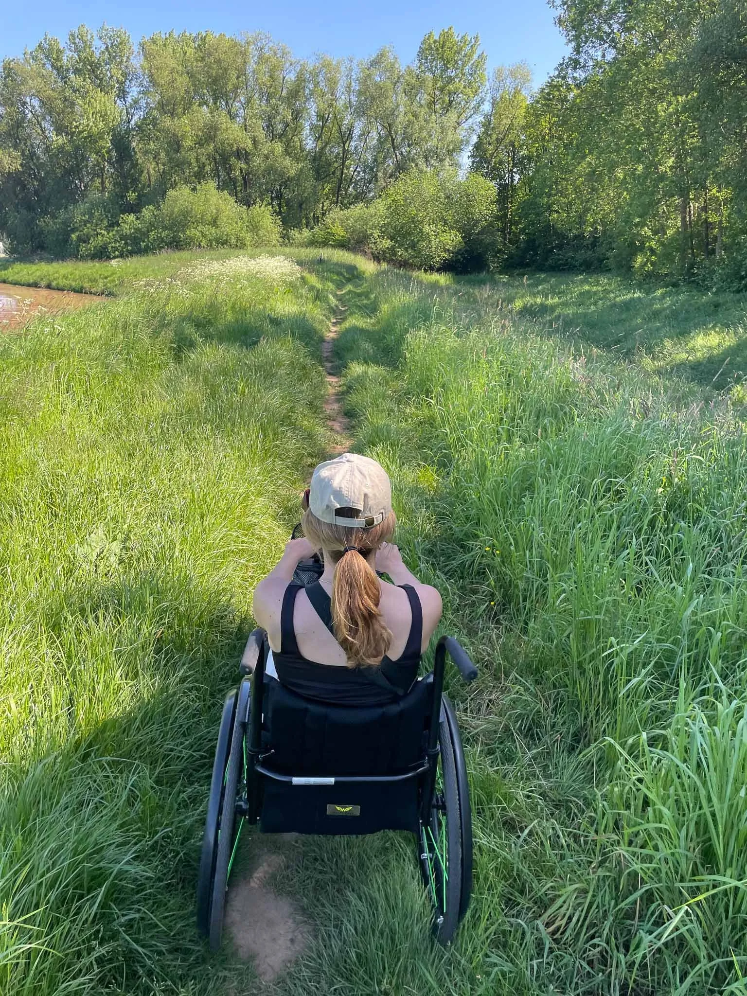 Een vrouw in een rolstoel wandelt door een groen gebied met gras en bomen onder een blauwe hemel.