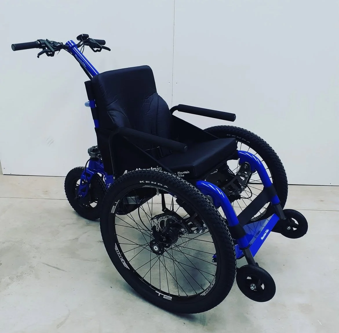 Mountain Trike e-Push. Een kinderrolstoel met een blauwe frame en zwarte zitplaats, geplaatst op een lichte vloer tegen een witte muur.