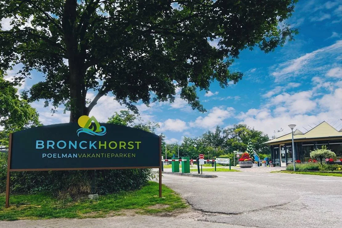Toegankelijke accommodatie aan het Pieterpad. Ingang van een vakantiepark met een bord dat BRONKHORST POELMAN VAKANTIEPARKEN aangeeft, een boom, een gebouw, een parkeerpoort en een lantaarnpaal onder een heldere blauwe lucht met wat wolken.