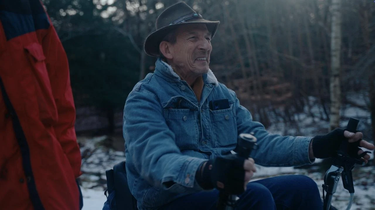 Mannen op een scooter in een bos, met een raam in de achtergrond, draagt een hoed en een denim jas.
