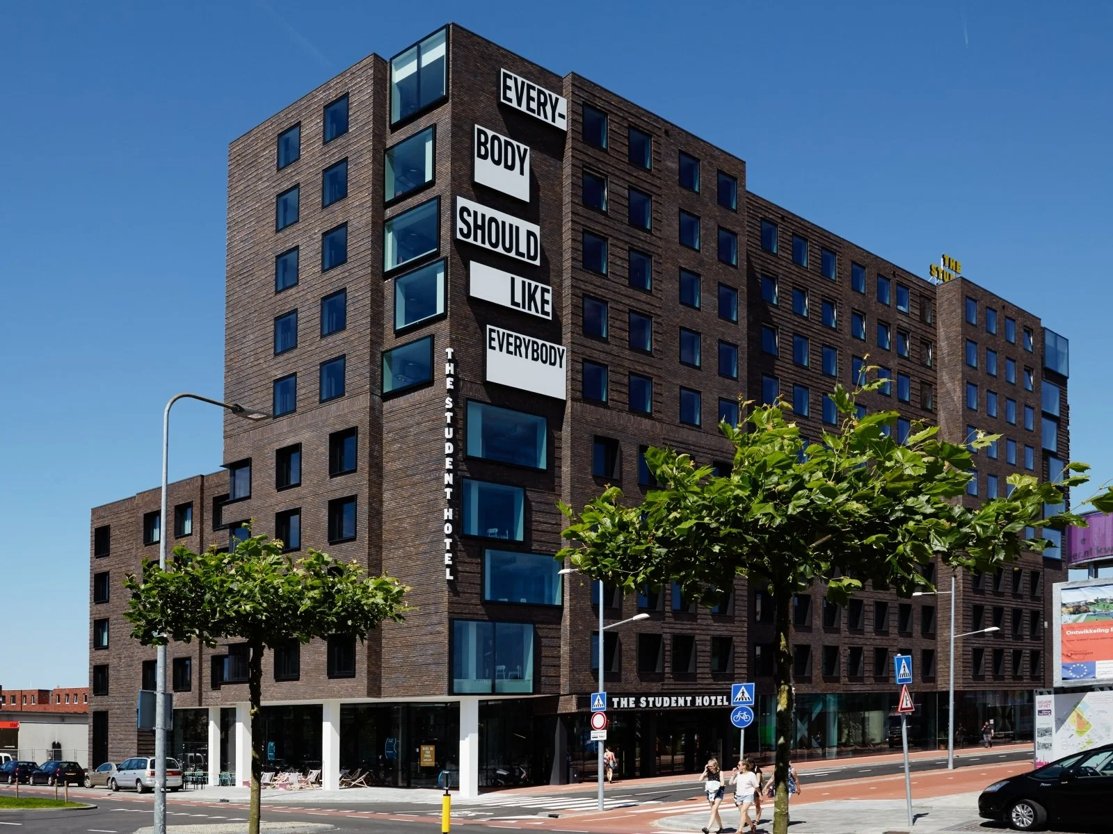 Een modern, bruin gekleurd hotelgebouw genaamd "The Student Hotel" met grote witte tekst op de gevel die zegt "Everybody should like everybody". Op straat voor het hotel lopen mensen en staan auto's geparkeerd.