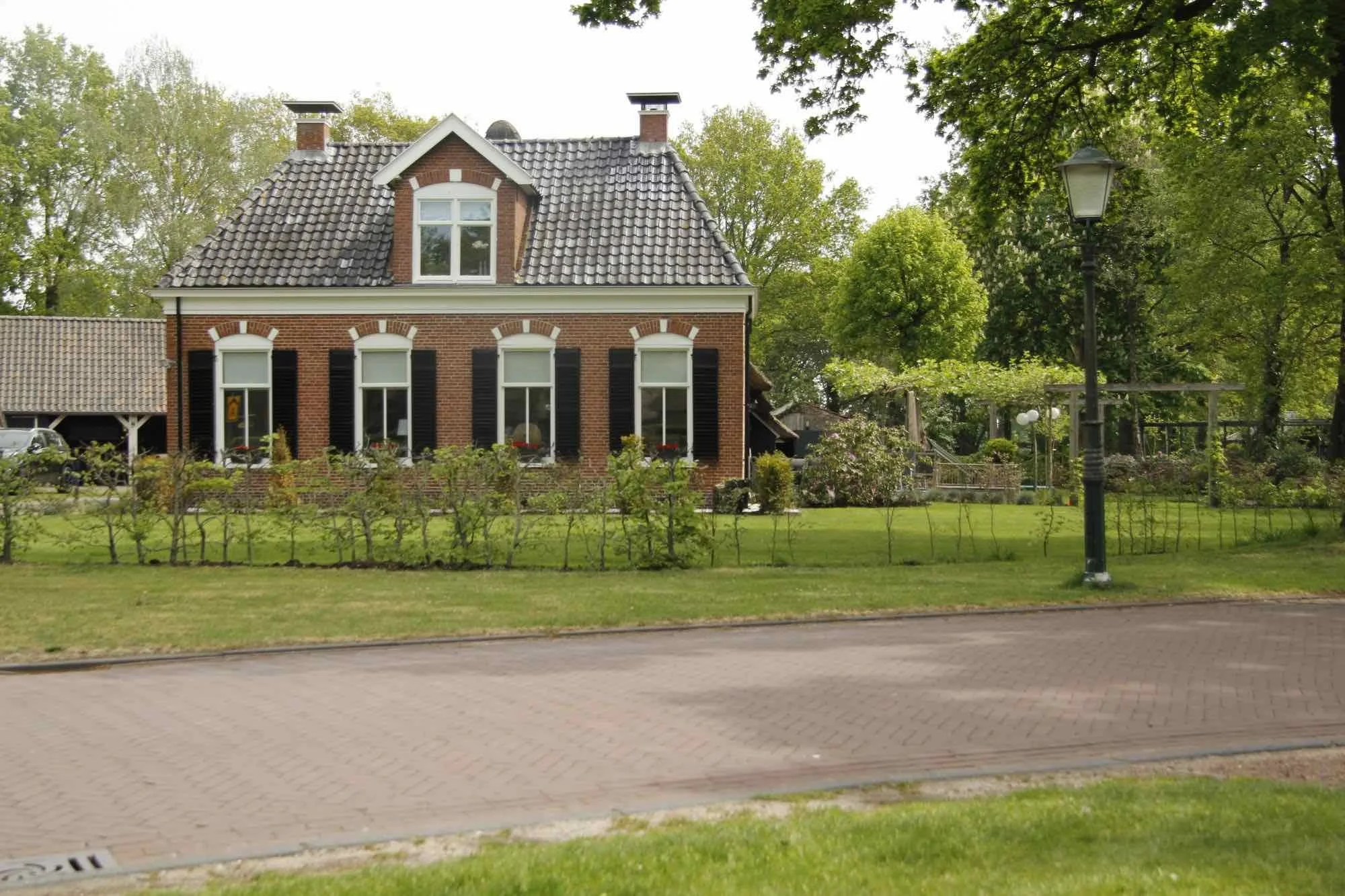 Een traditionele Nederlandse brick house met een tuin en bomen, een straat en een lantaarnpaal op de voorgrond.
