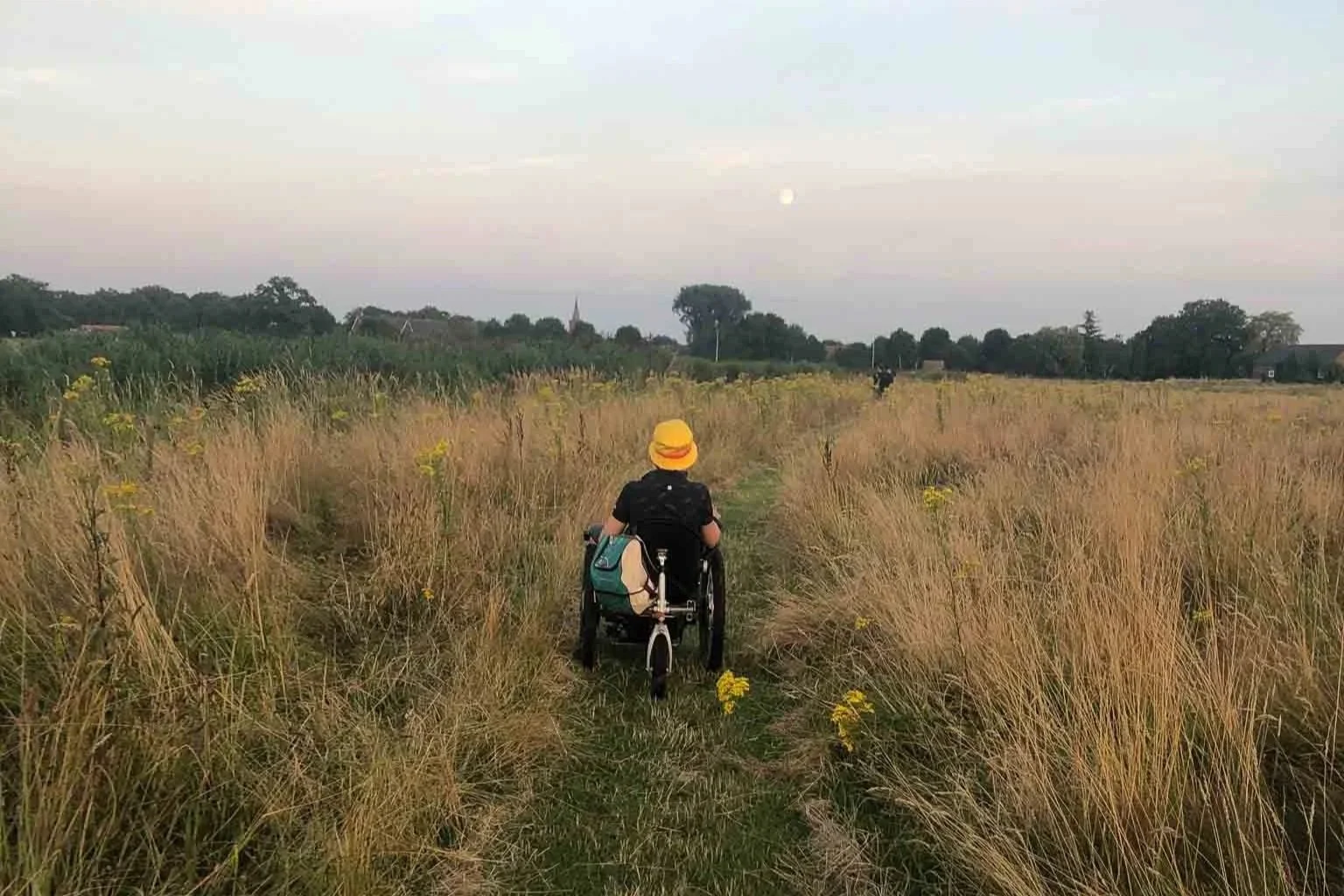 Persoon in een rolstoel fietst door een veld met hoog gras en bloemen, onder een ciel met de maan zichtbaar op de achtergrond.