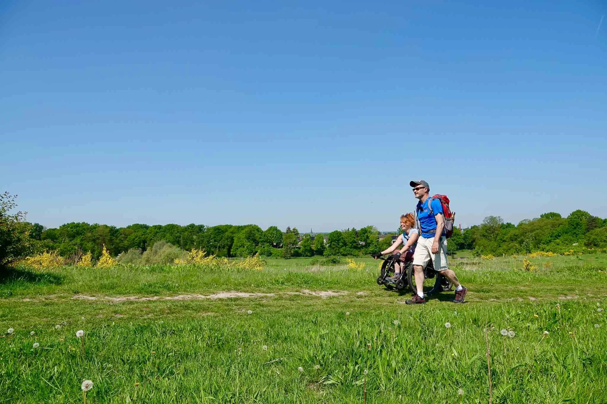 Twee mensen wandelen in een groen veld onder een heldere blauwe lucht. Een man en een jonge meisje, beide dragen sportkleding en backpacks, genieten van een dag buiten.
