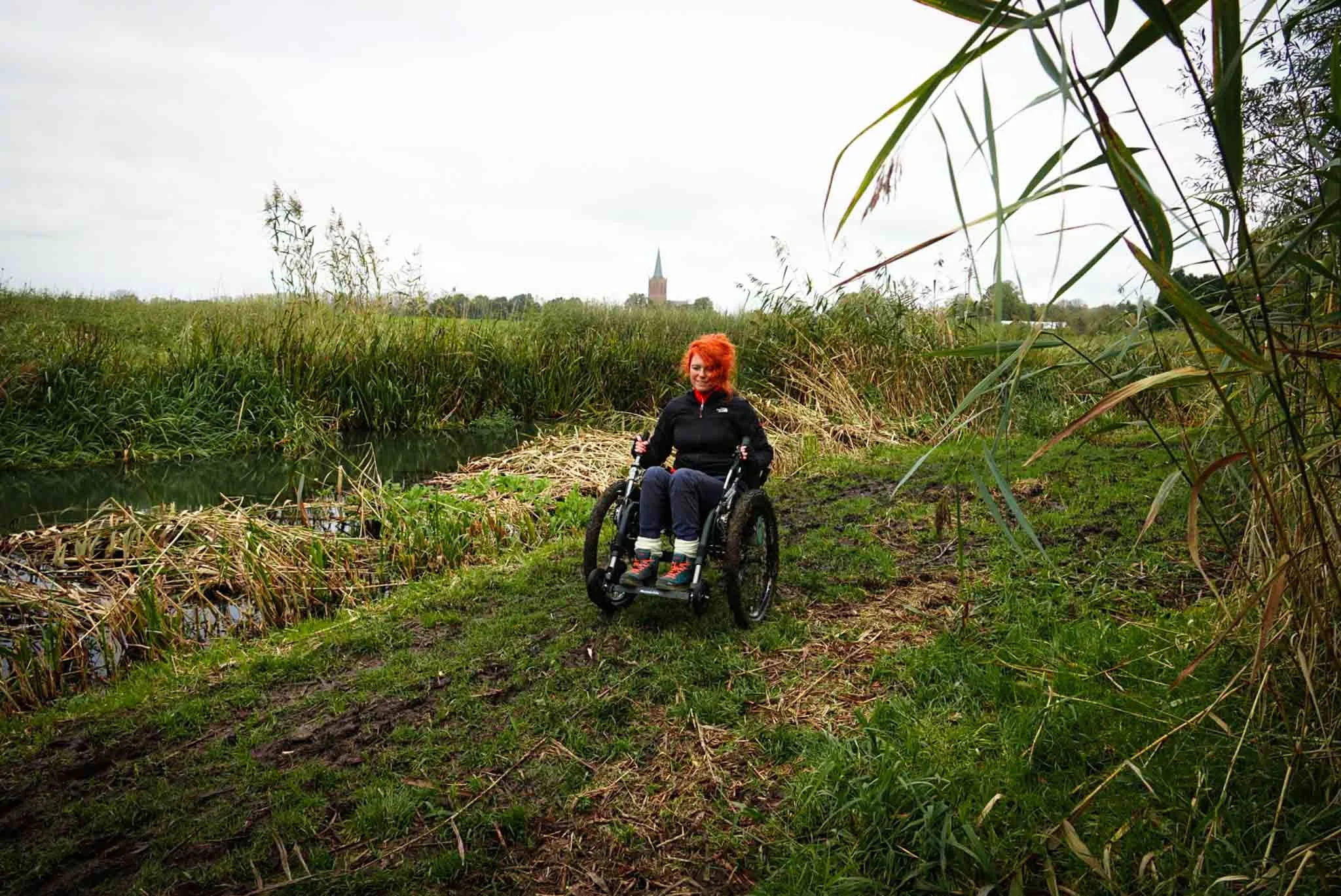 Vrouw in rolstoel op een grasachtig pad langs een waterkant, met groene rietvelden en een kerk in de achtergrond.