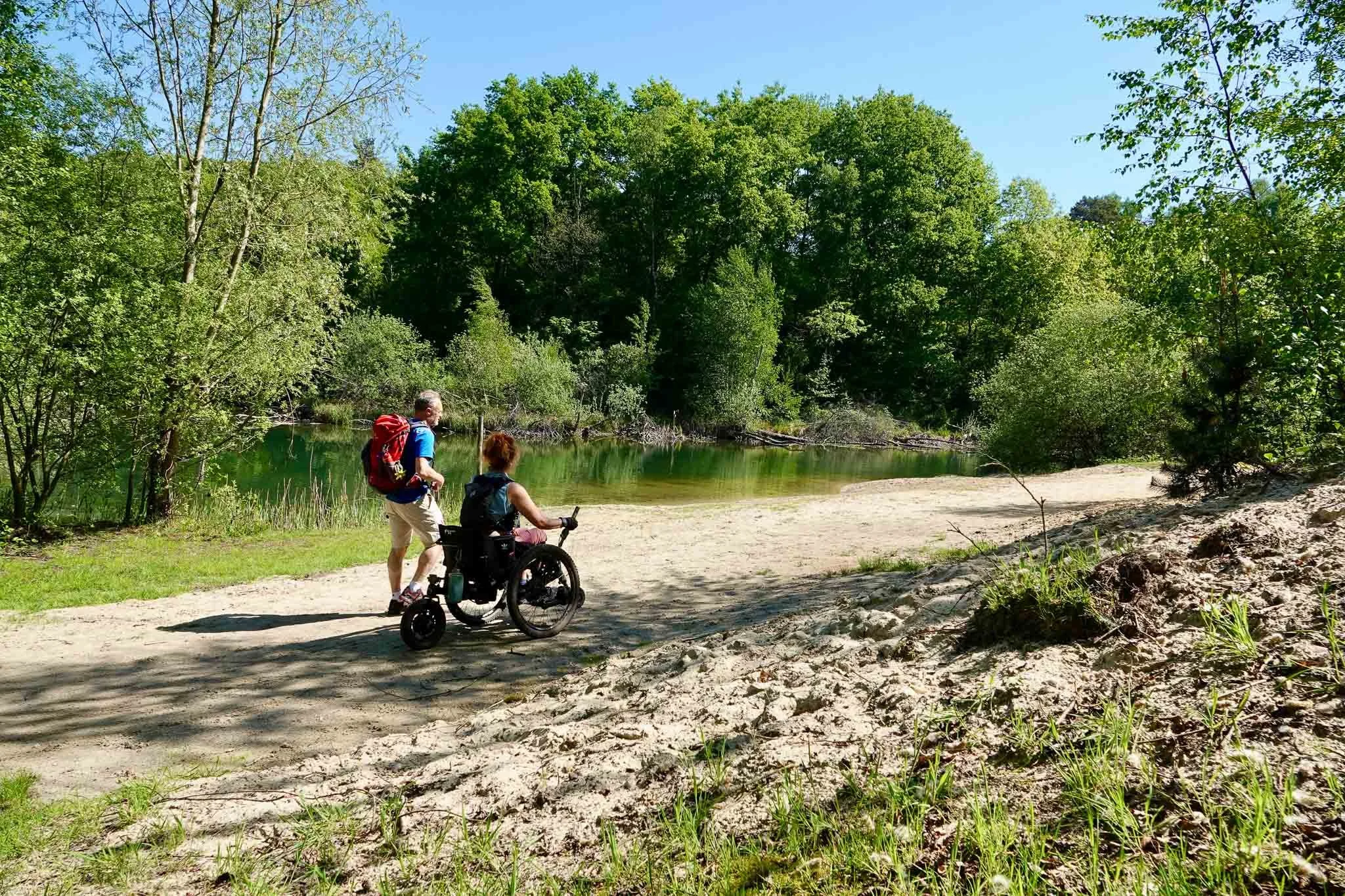 Twee mensen, een man en een vrouw in een rolstoel, wandelen langs een rivier in een groen bos op een zonnige dag.