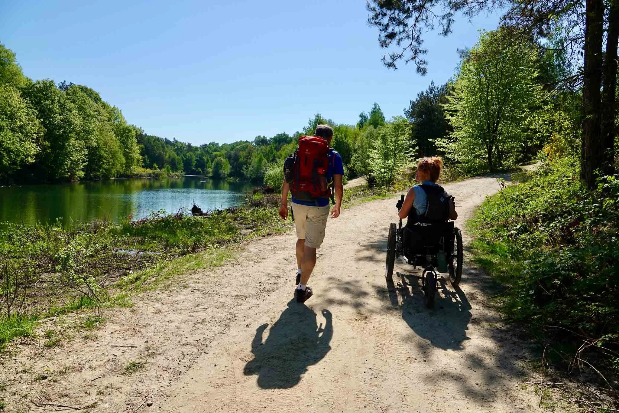 Twee mensen wandelen langs een rivier in een bosrijke omgeving op een zonnige dag, een persoon in een rolstoel en een persoon met rugzak.