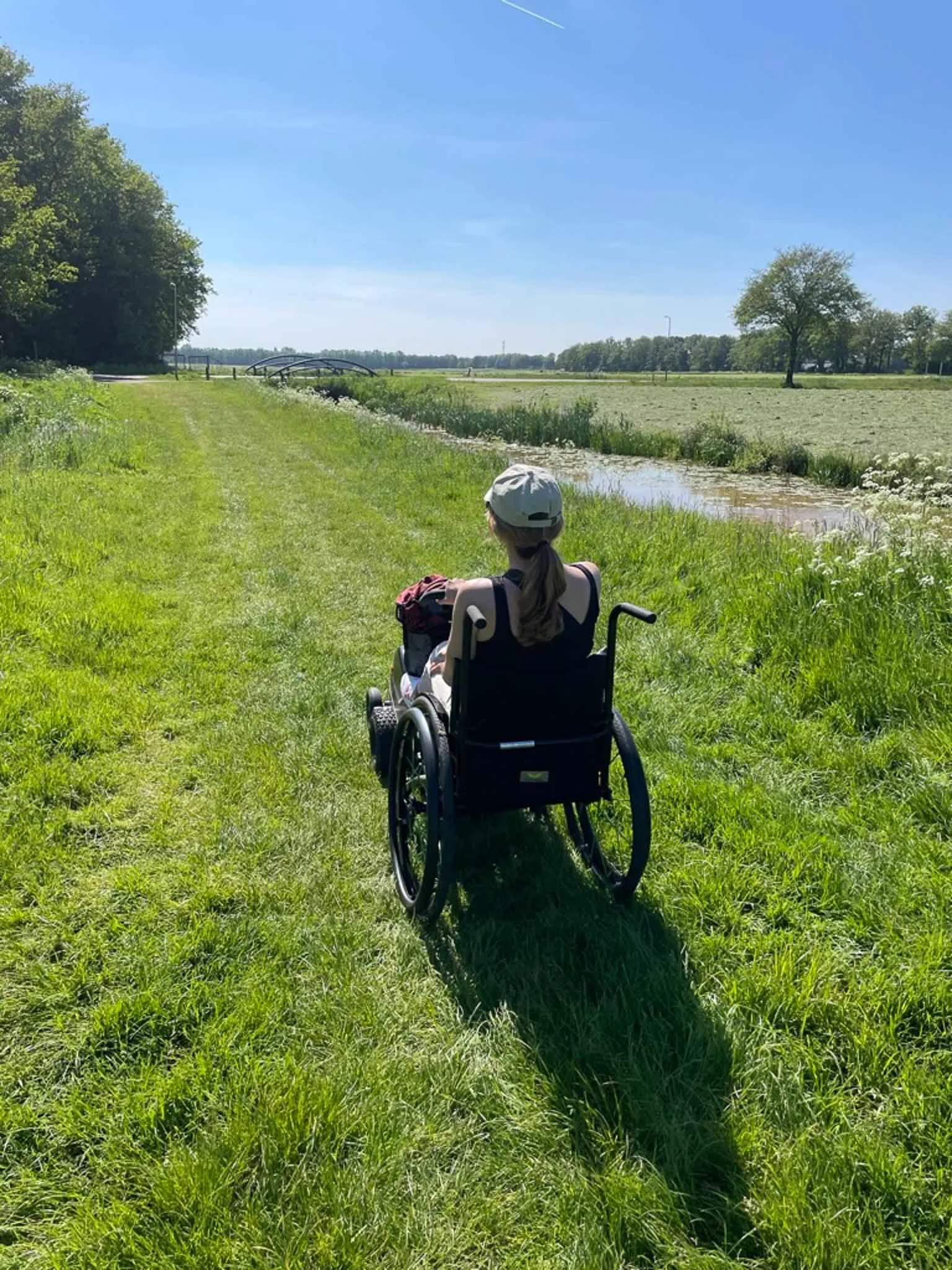 Een persoon in een rolstoel wandelt over een grasstrook langs een kanaal onder een blauwe hemel.