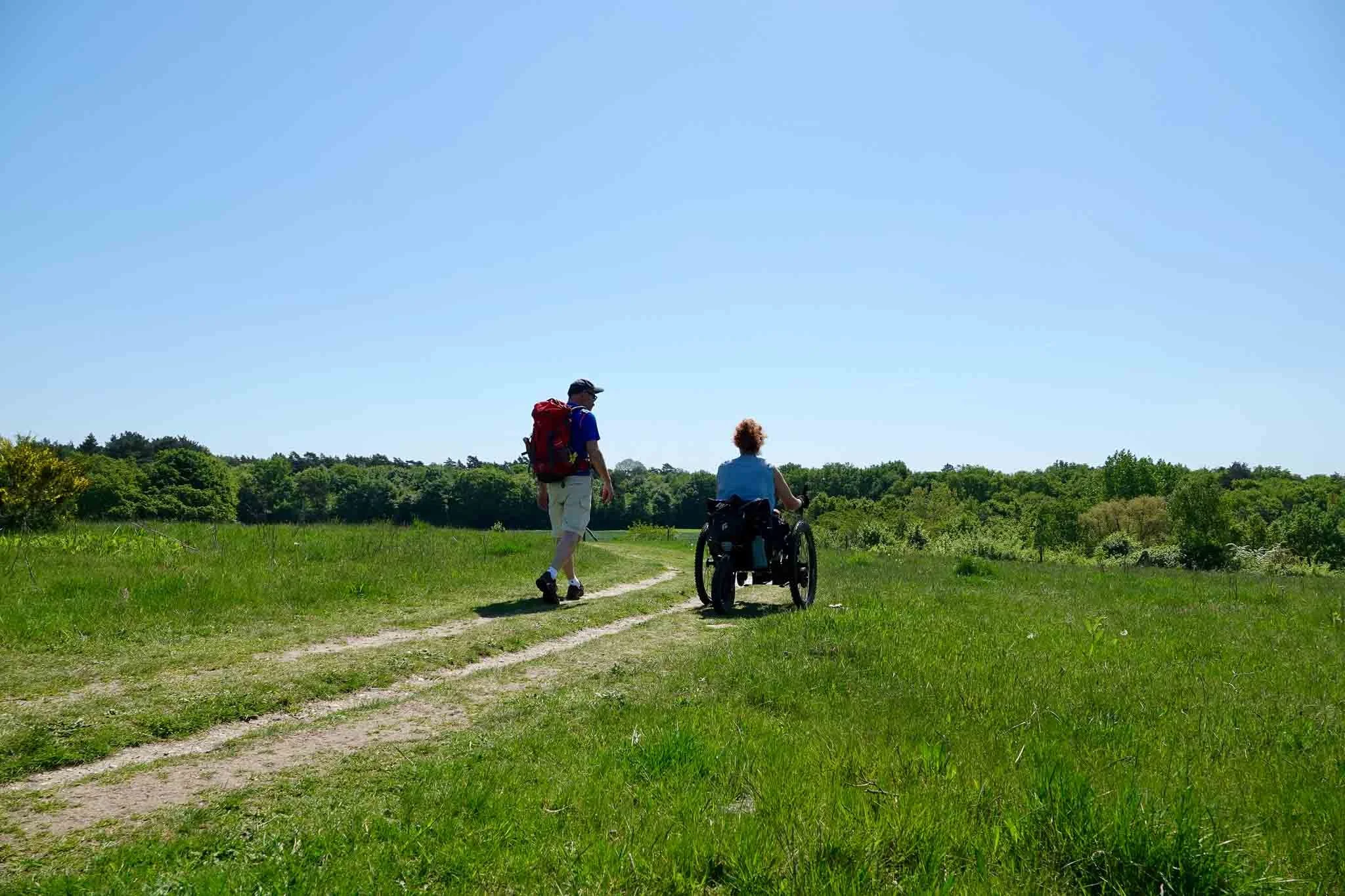 Twee mensen wandelen op een bospad in een uitgestrekt groene landschap onder een heldere blauwe lucht, één persoon zit in een rolstoel en de ander loopt naast hen.