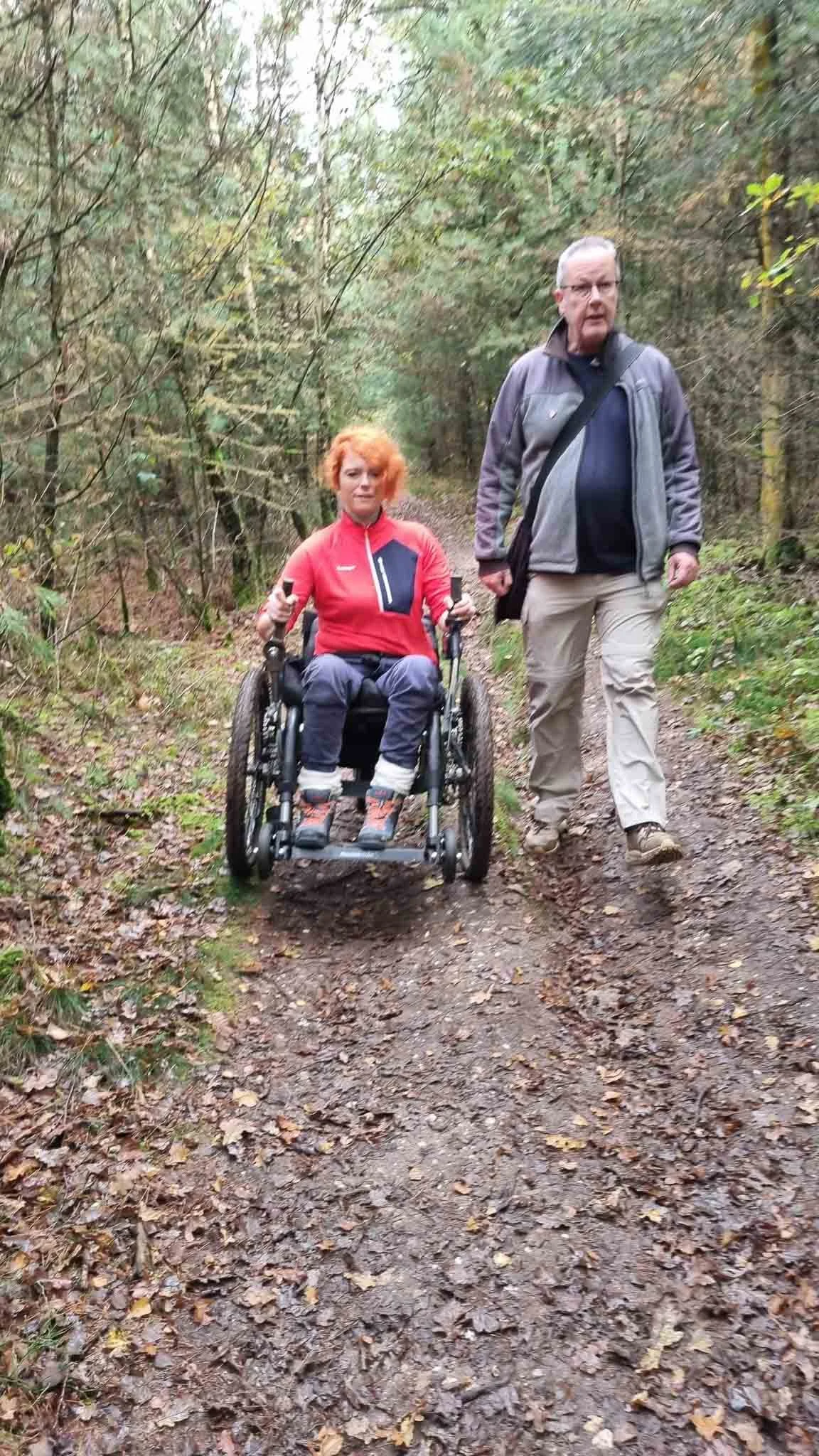 Man en vrouw wandelen in het bos. De vrouw zit in een rolstoel. Ze lijken samen te genieten van een wandeling op een modderig pad.