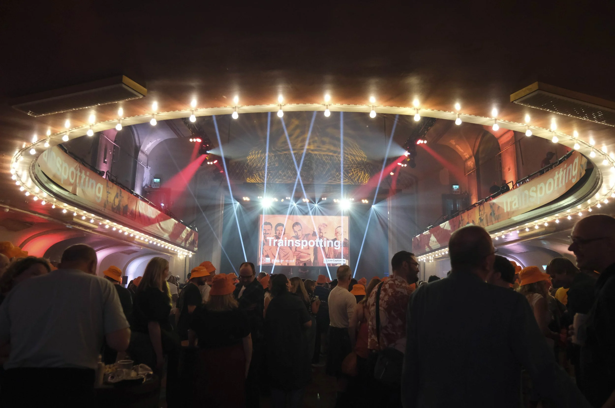 Crowd gathered in a busy auditorium with orange lights and Trainspotting branding. Some audience members wearing orange bucket hats.