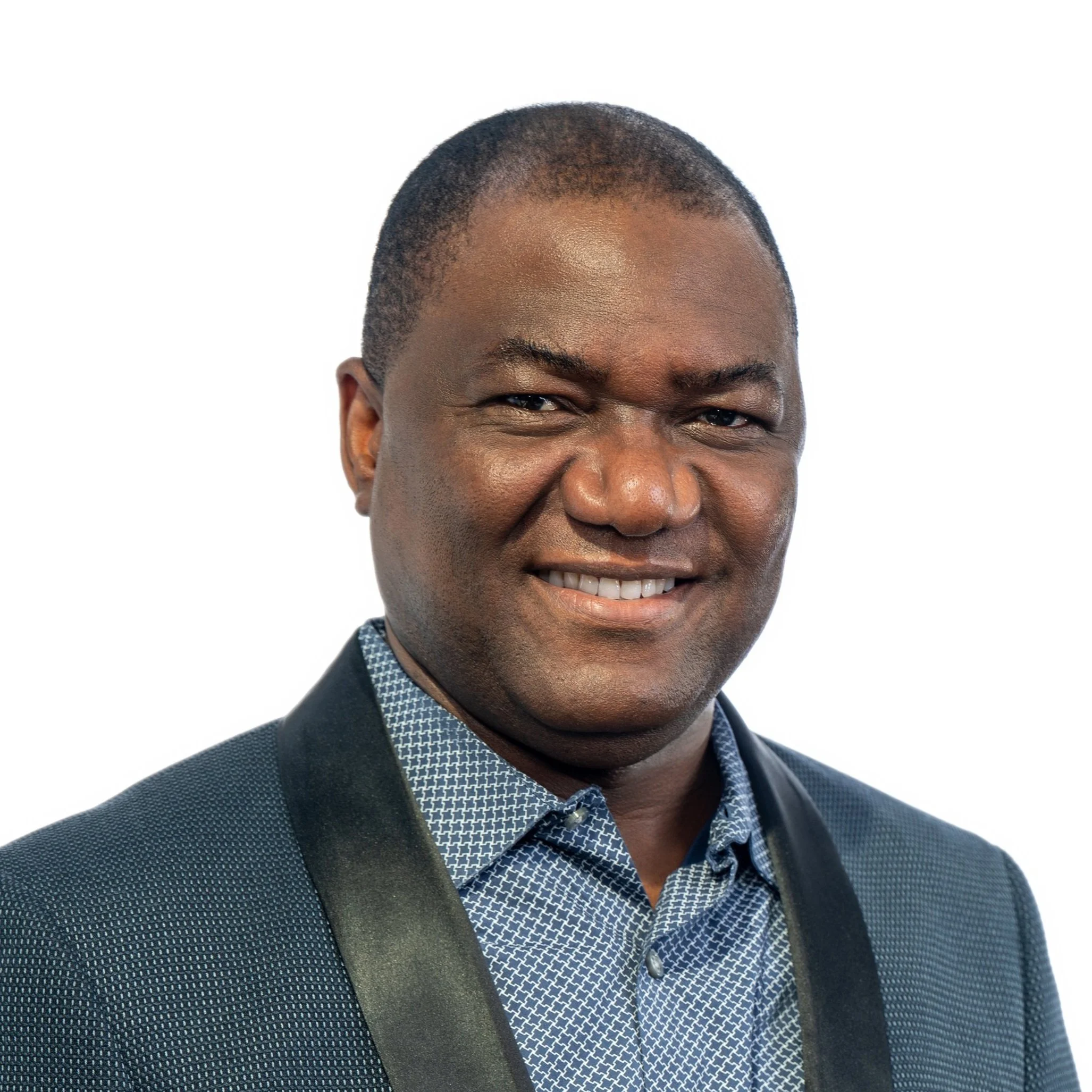 Headshot of a smiling African American man in a patterned blue shirt and dark blazer with a solid white background.
