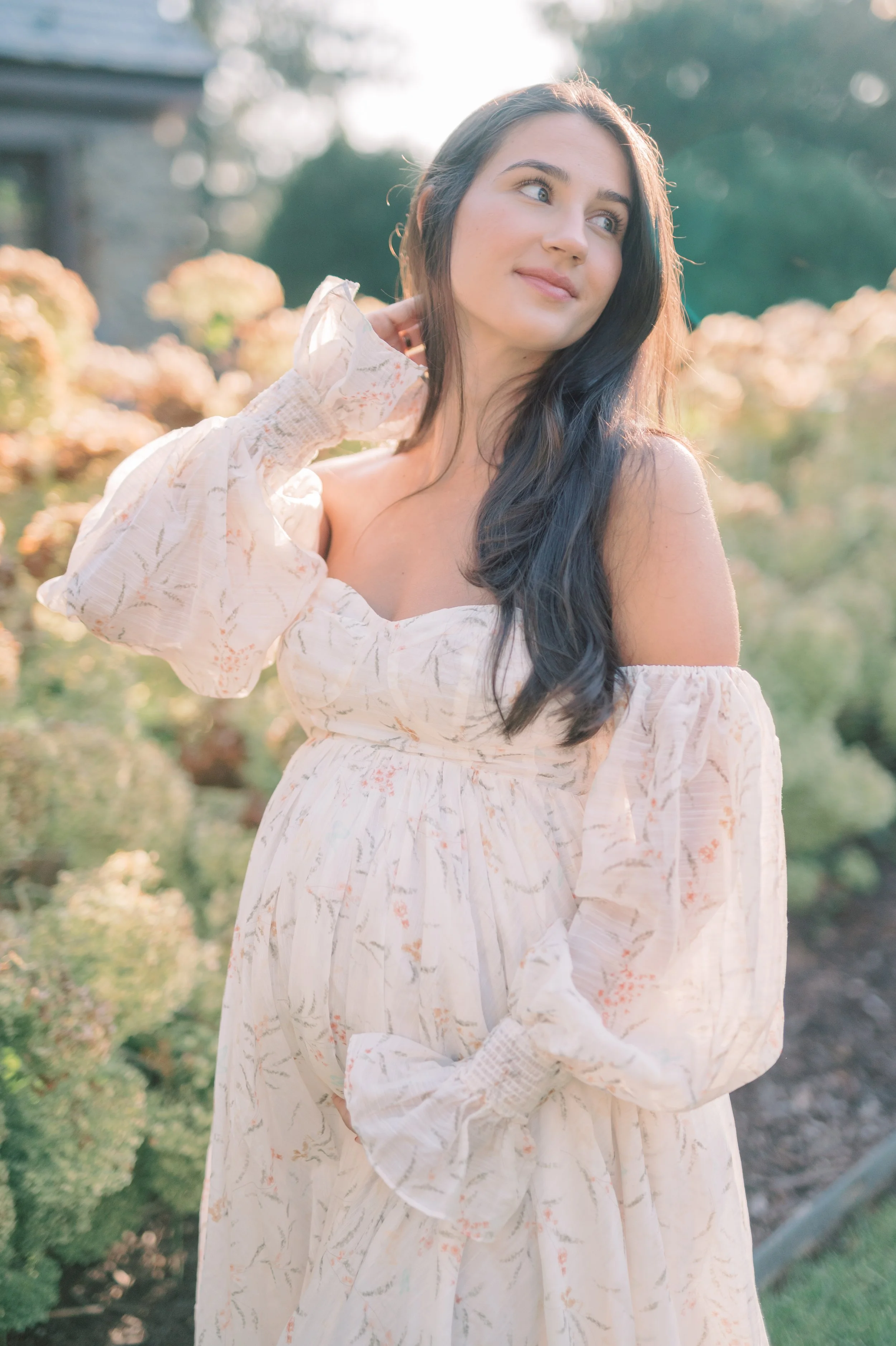 A woman with long dark hair wearing a flowing, off-shoulder cream dress with pink and green patterns, standing outdoors with blurred bushes and flowers in the background, touching her neck with one hand and looking to the side.