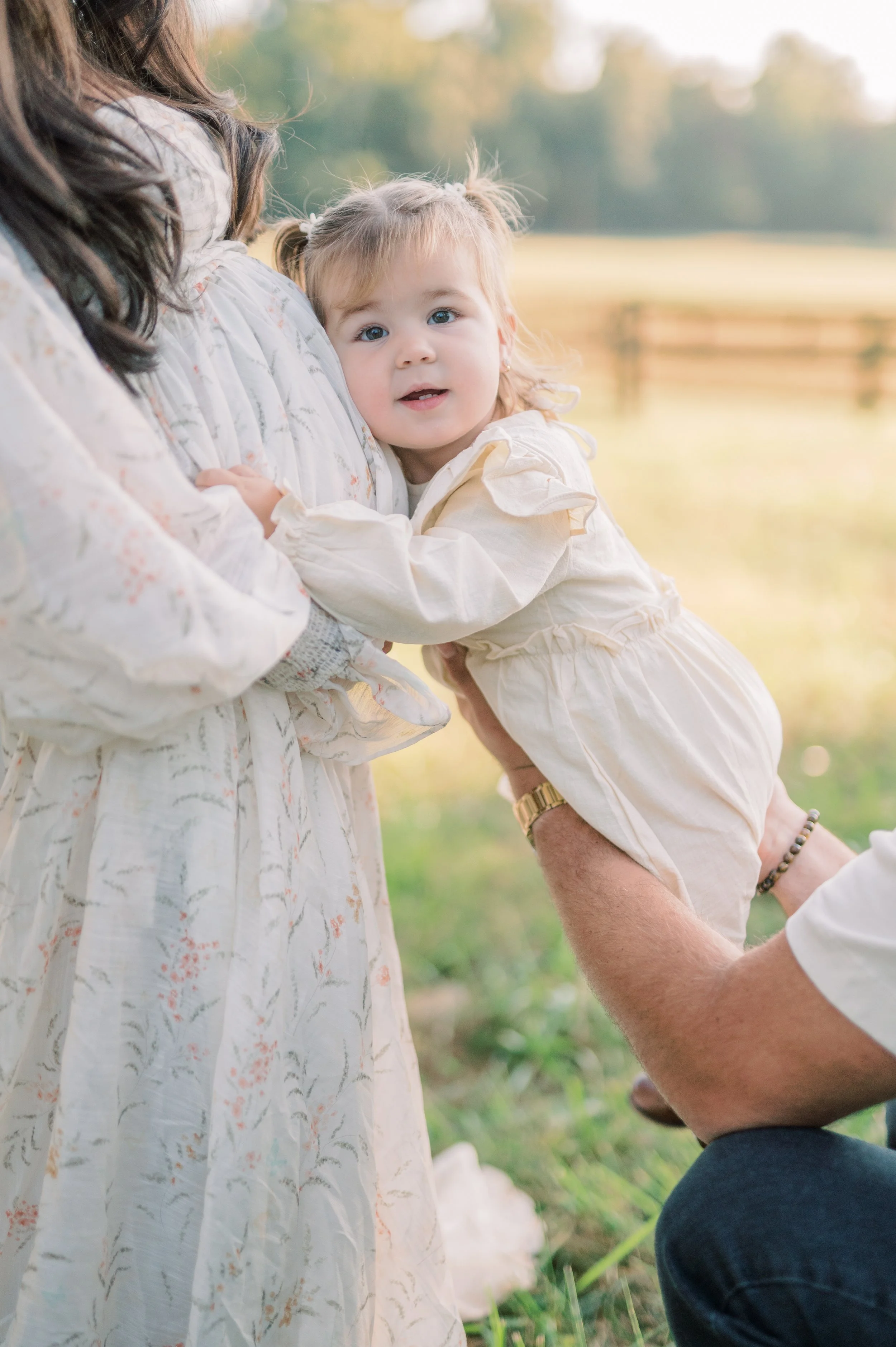 A young girl in a cream-colored dress being held by an adult man with a gold watch, while an adult woman in a patterned dress holds her. They are outdoors in a grassy area with trees in the background, during late afternoon or early evening.