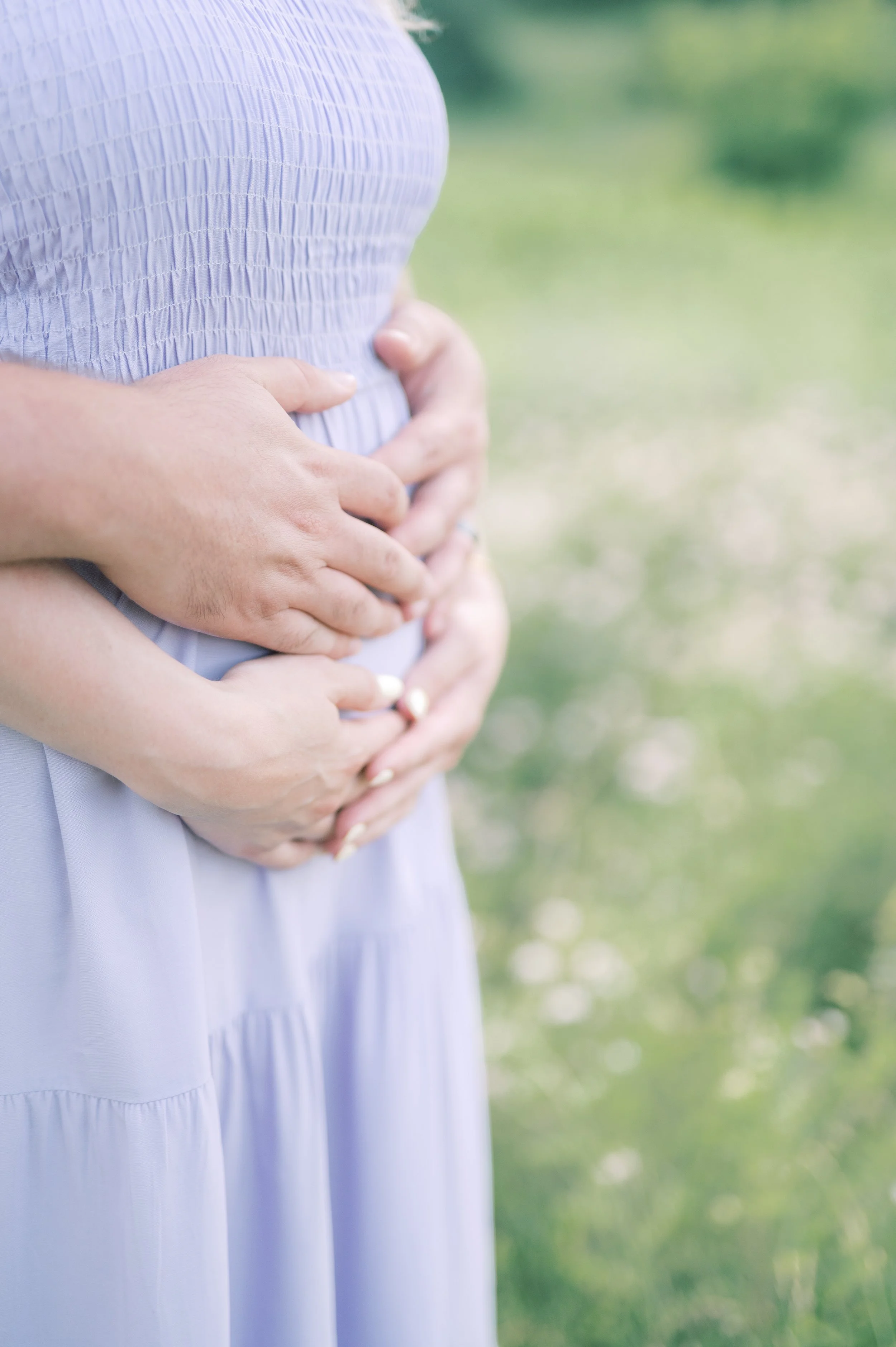 Two women with intertwined hands are clutching a pregnant belly, outdoors on a grassy field.