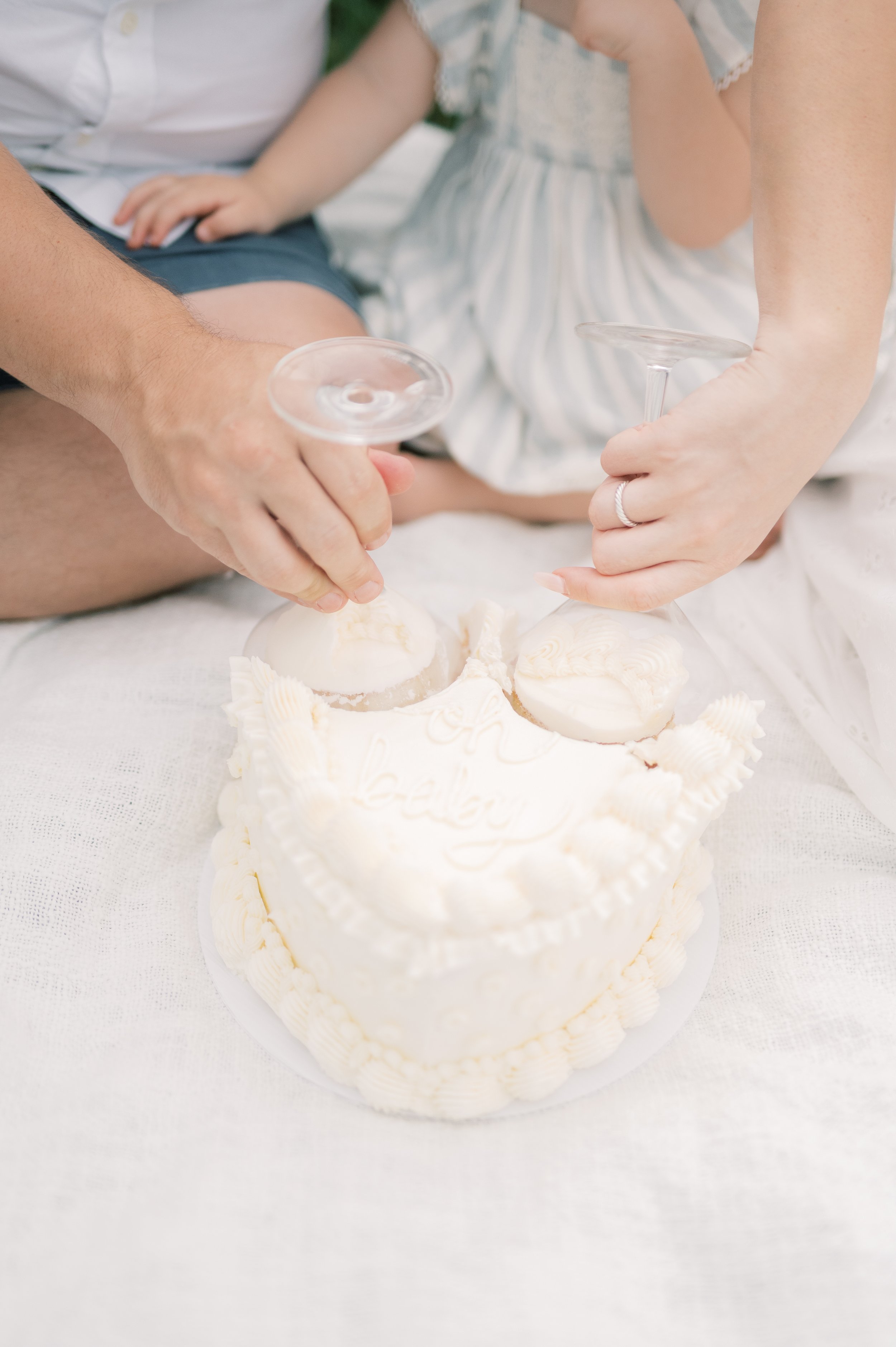Close-up of a white birthday cake with two small round desserts, people holding empty glasses for a toast, celebrating with a cake on a white table.