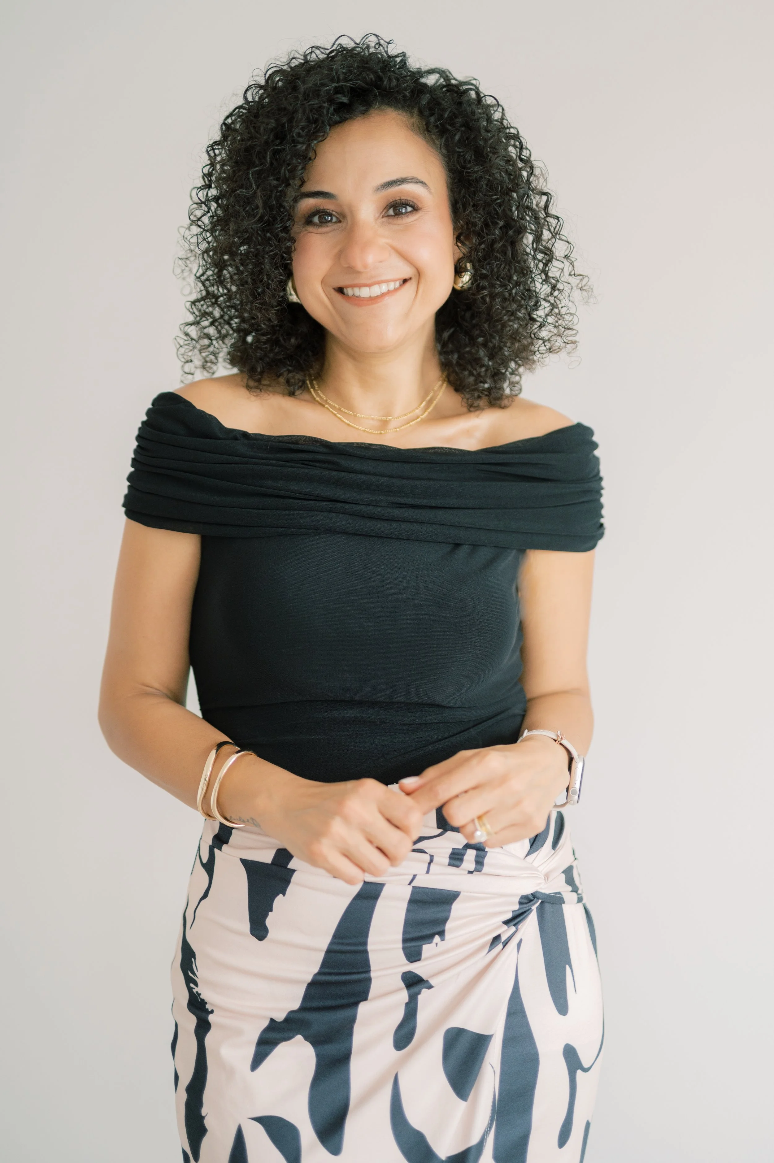 A woman with curly black hair smiling, wearing a black off-the-shoulder top and patterned skirt, standing against a plain white background.