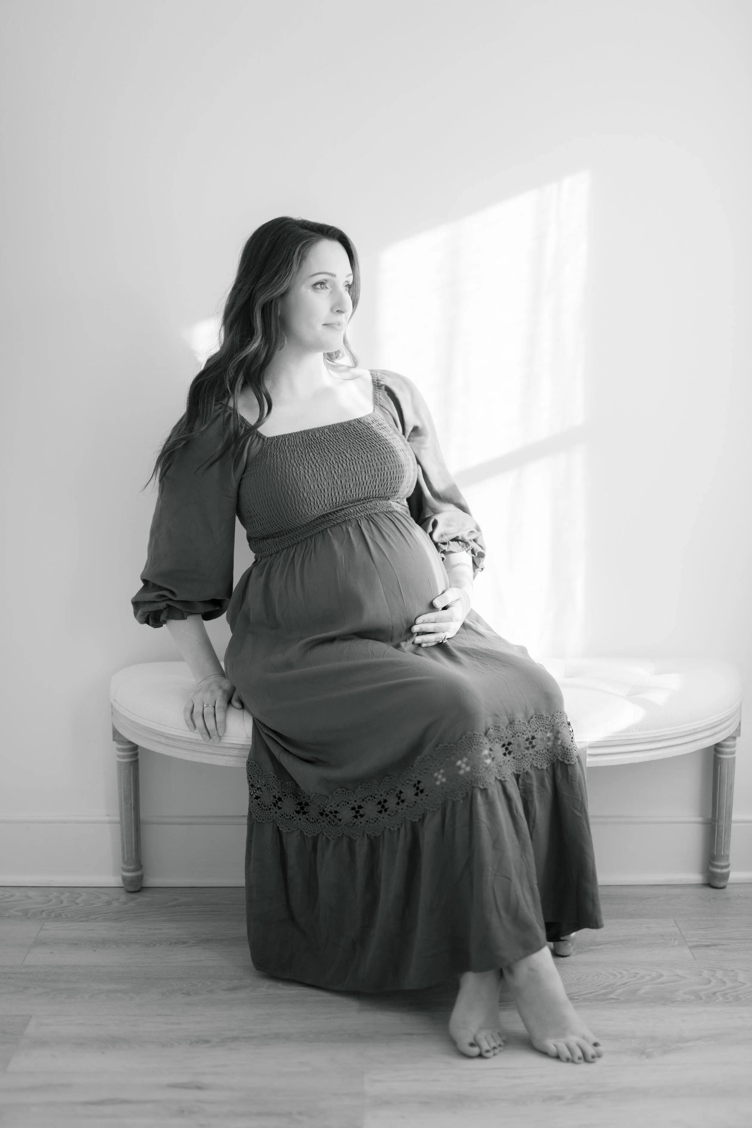 A pregnant woman sitting on a bench, looking to the side, with sunlight casting shadows on the wall behind her.